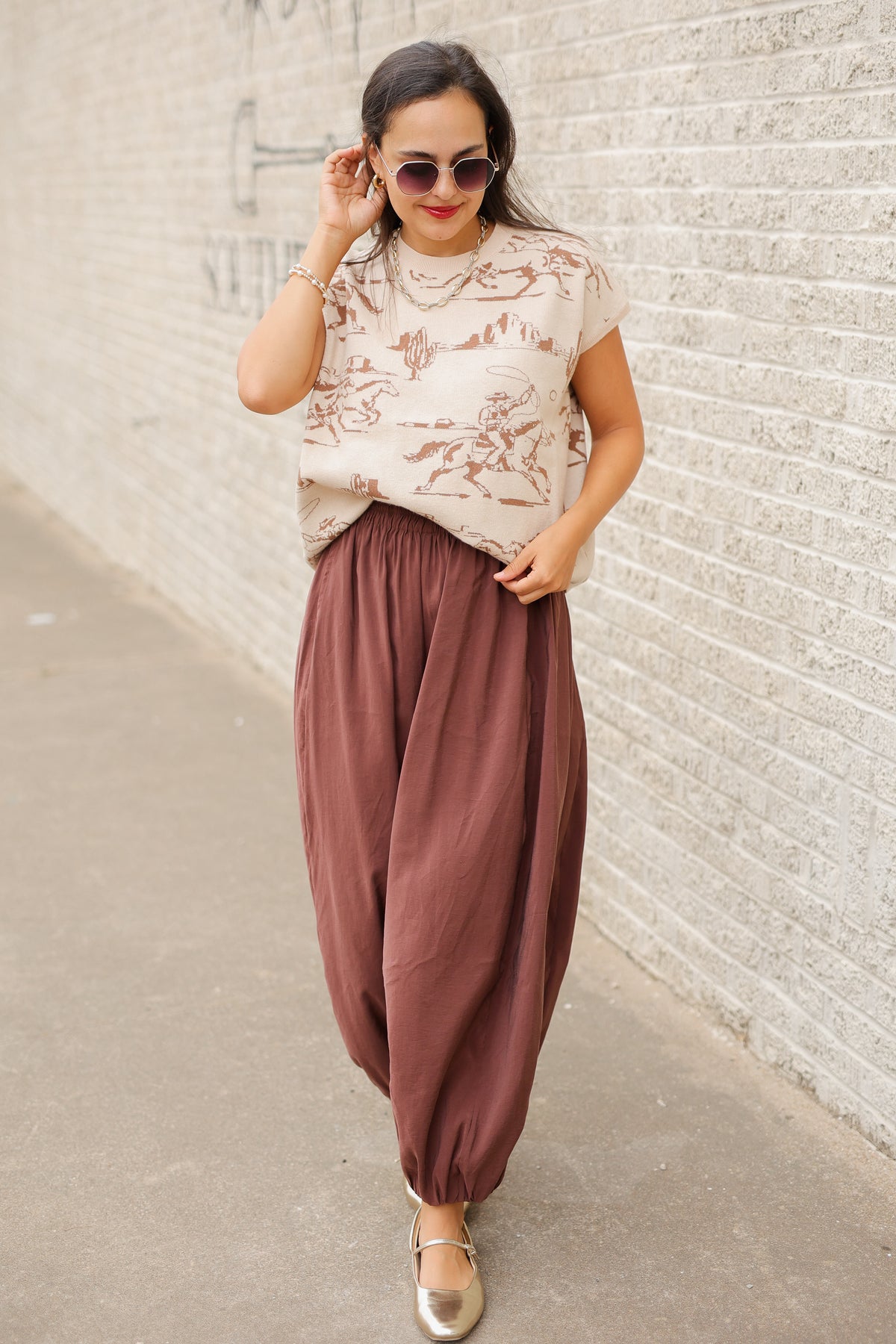 Model wearing Bronco Trail Knit Top in Ecru featuring taupe western cowboy, bucking horse, and desert cactus illustrations, styled with the Tulara Barrel Pants – Brown from Southern House Clothing.