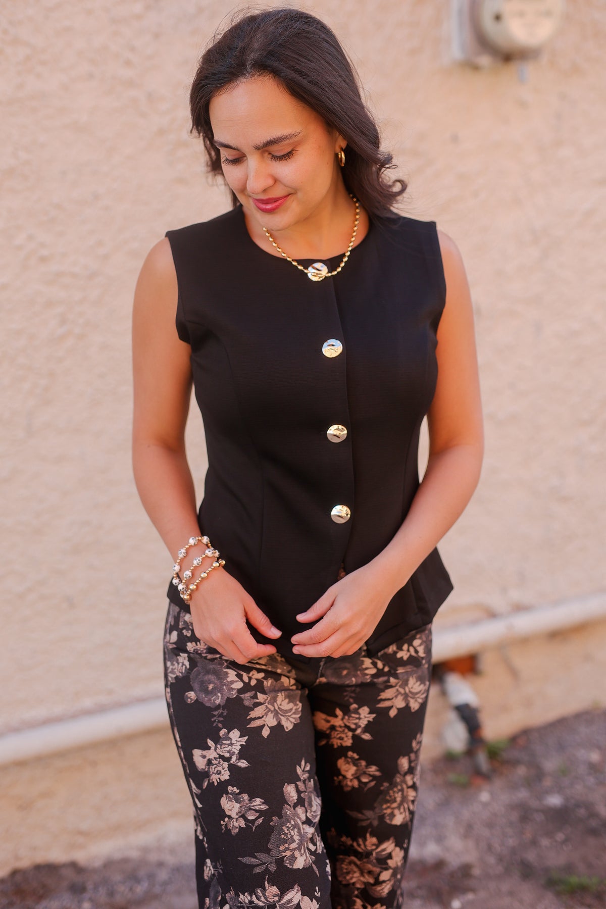 Woman wearing a black sleeveless top with gold buttons and floral pants against a beige wall.