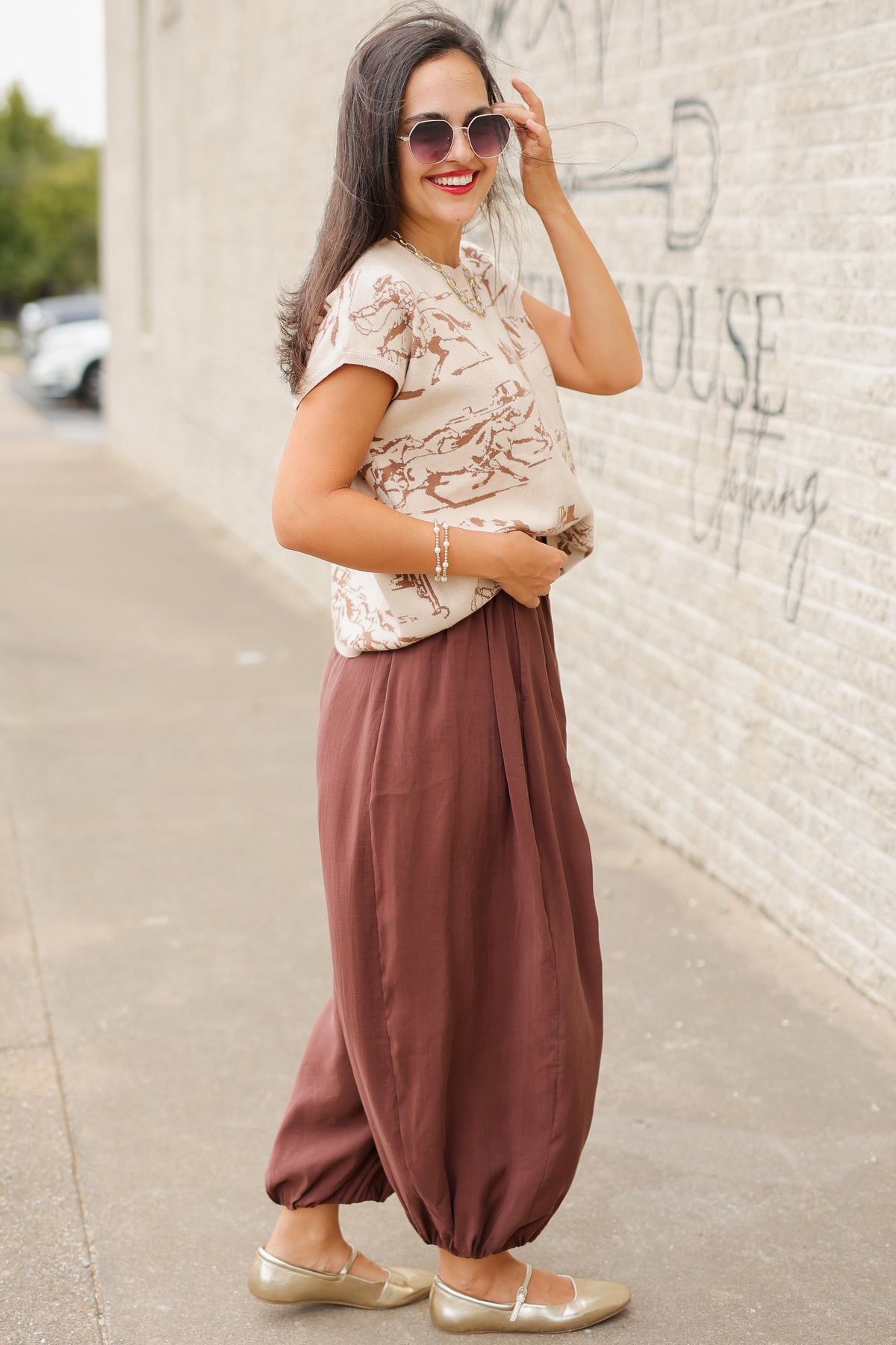 Model wearing Bronco Trail Knit Top in Ecru featuring taupe western cowboy, bucking horse, and desert cactus illustrations, styled with the Tulara Barrel Pants – Brown from Southern House Clothing.