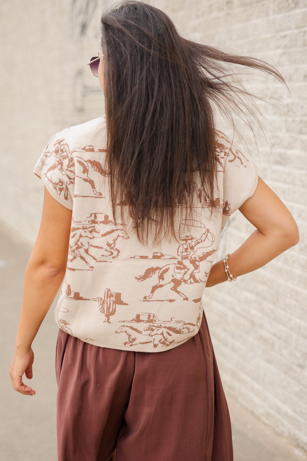 Back view of a model wearing Bronco Trail Knit Top in Ecru featuring taupe western cowboy, bucking horse, and desert cactus illustrations, styled with the Tulara Barrel Pants – Brown from Southern House Clothing.