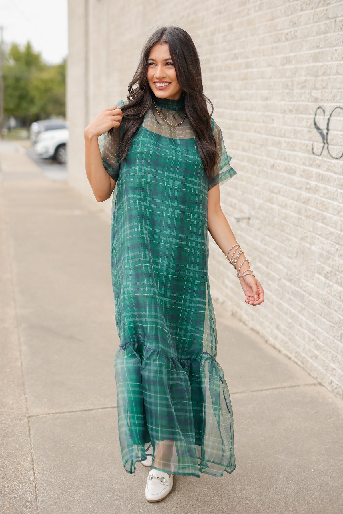 Model standing outside wearging the Glenhaven Plaid Ruffle Maxi Dress in emerald green and navy featuring a high neckline, sheer short sleeves, relaxed silhouette, and flowy ruffled hem. Southern House Clothing