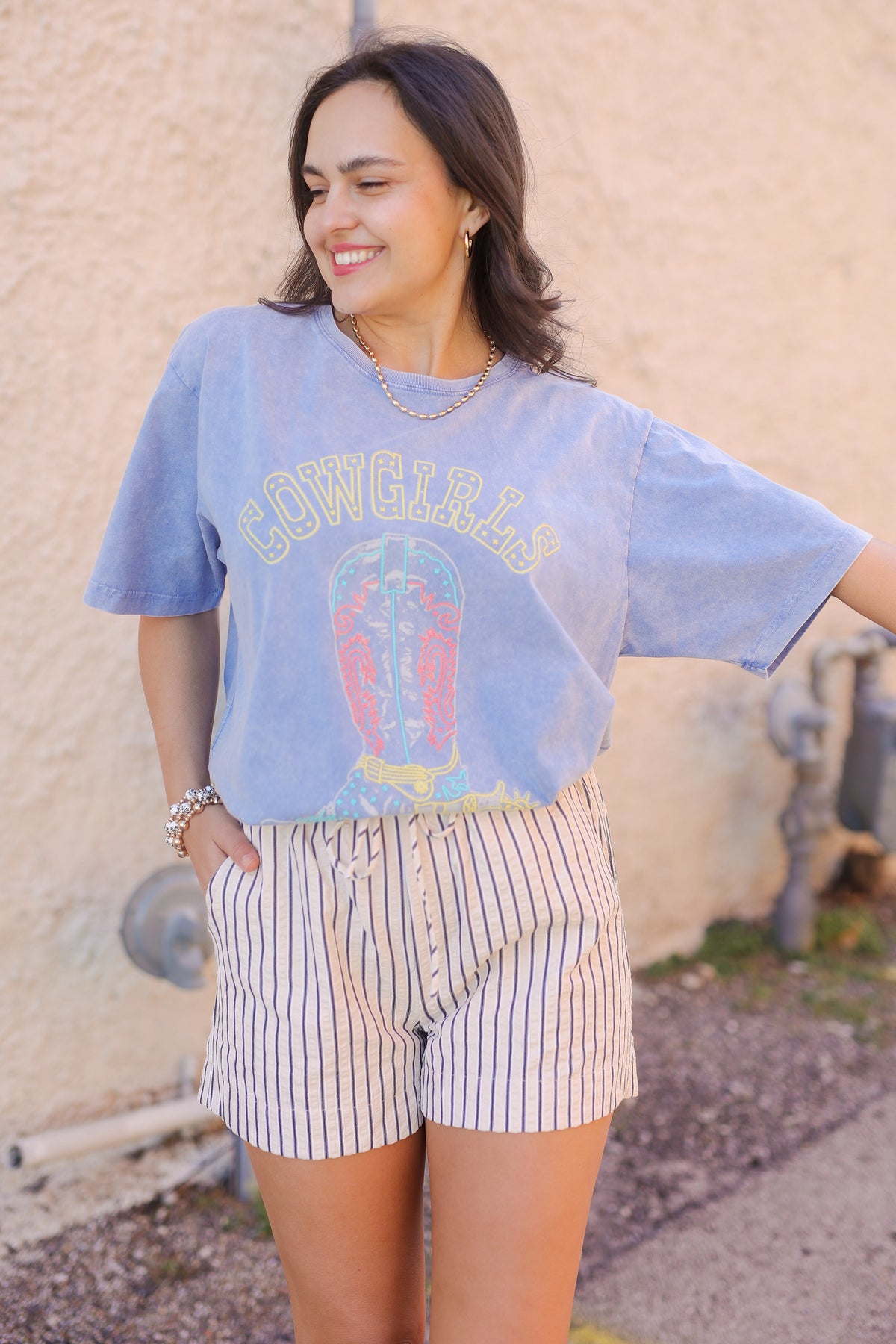 Woman wearing a light blue graphic t-shirt and striped shorts standing against a beige wall.