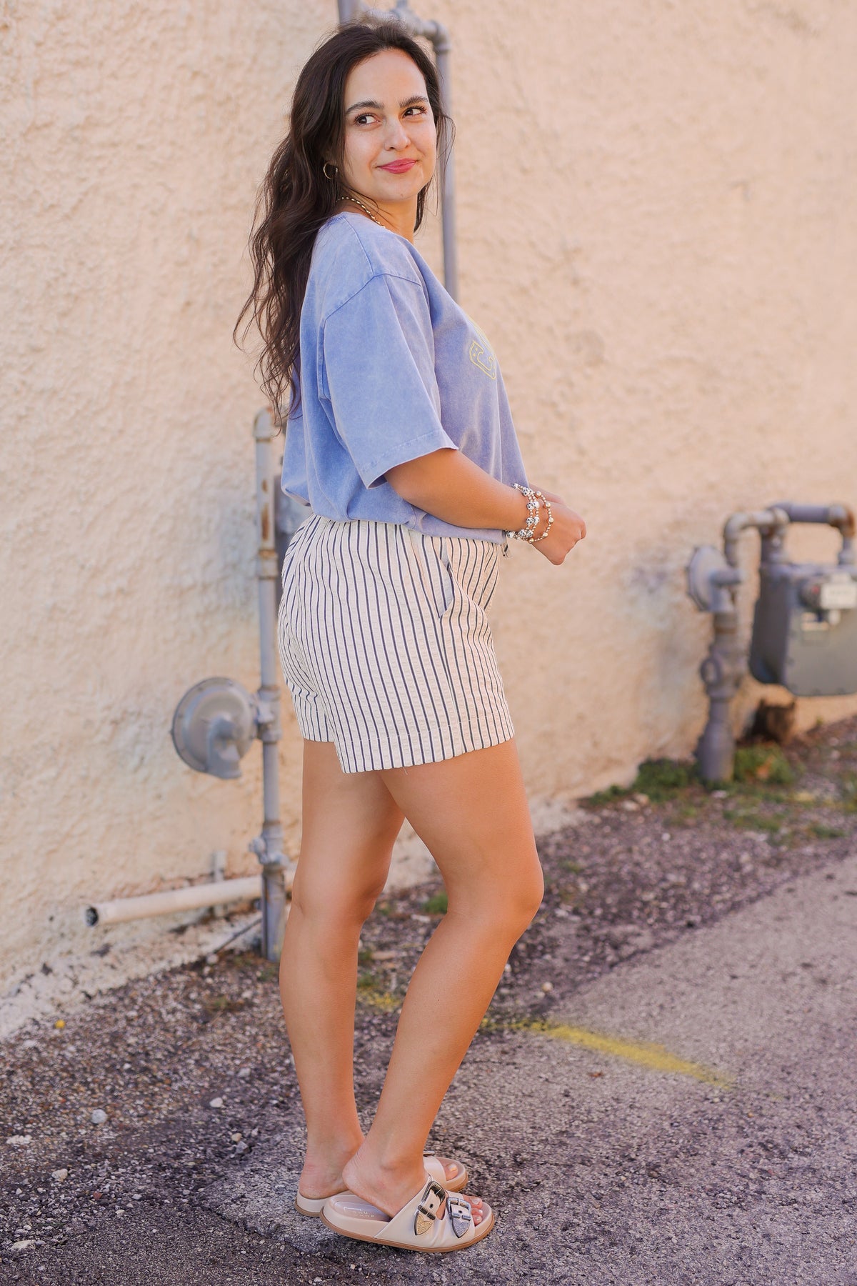 Woman in a light blue shirt and striped shorts standing against a beige wall with pipes.