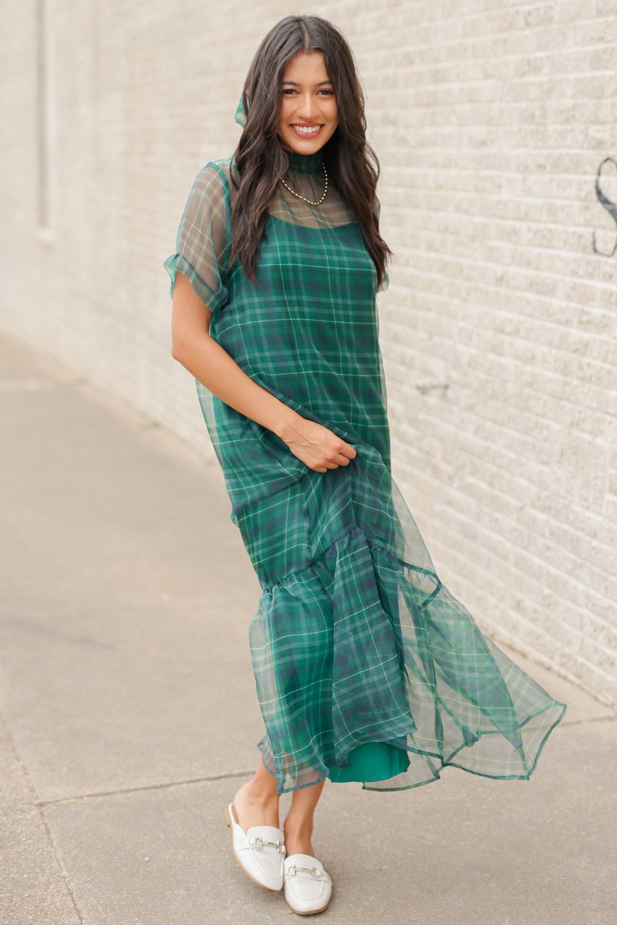 Model standing outside wearing the Glenhaven Plaid Ruffle Maxi Dress in emerald green and navy featuring a high neckline, sheer short sleeves, relaxed silhouette, and flowy ruffled hem. Southern House Clothing