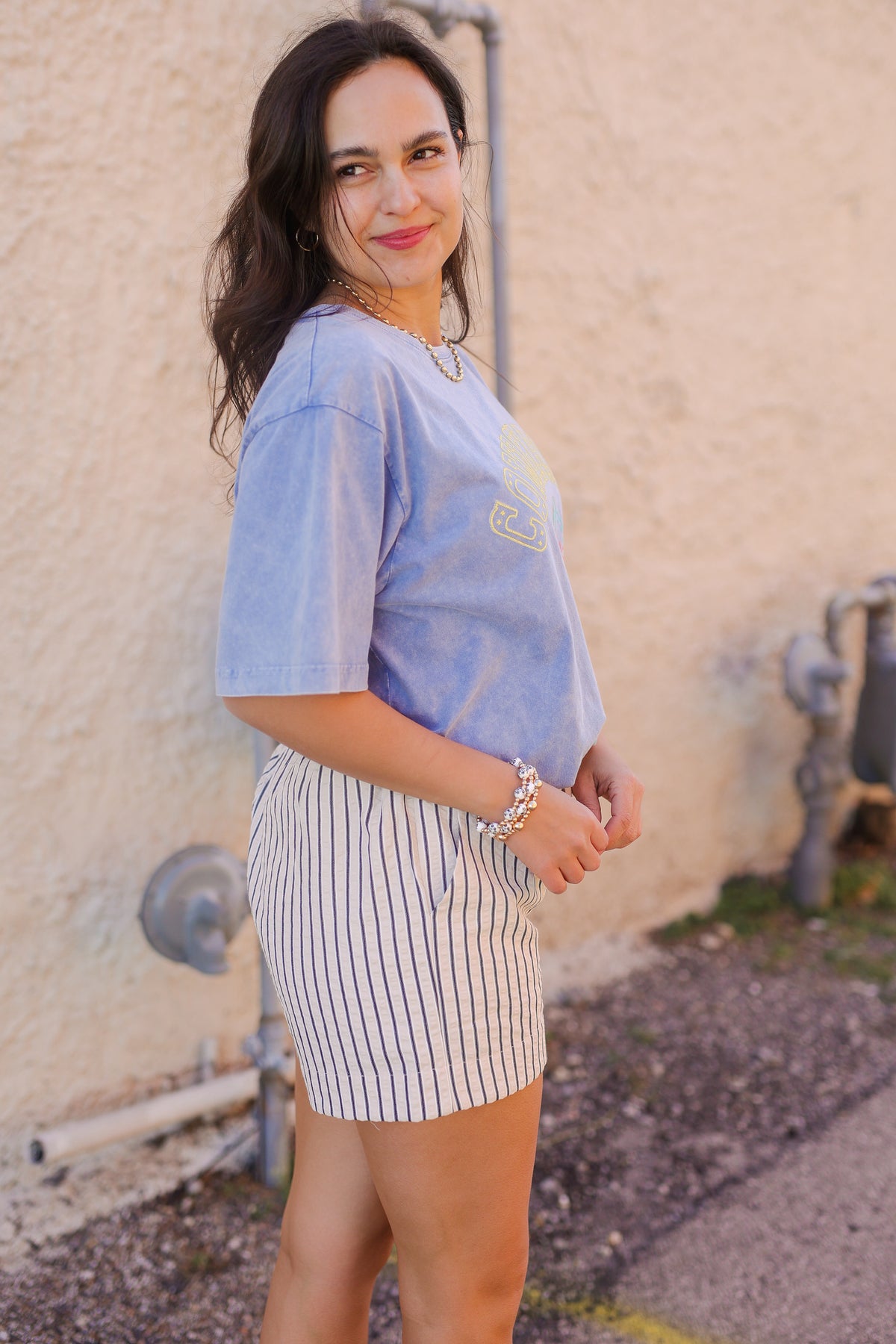 Woman wearing a light blue shirt and striped shorts standing against a beige wall.