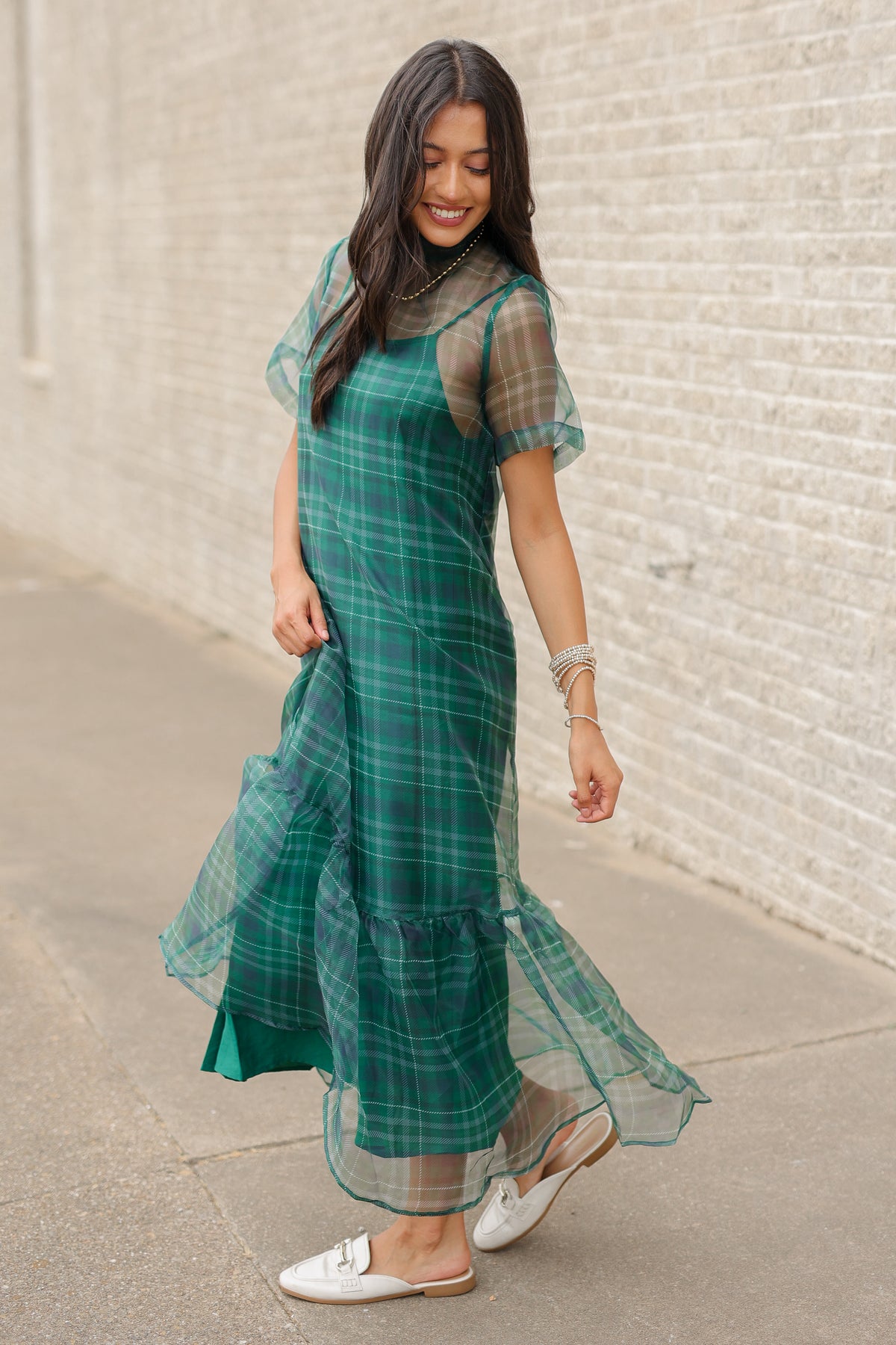 Model standing outside wearing the Glenhaven Plaid Ruffle Maxi Dress in emerald green and navy featuring a high neckline, sheer short sleeves, relaxed silhouette, and flowy ruffled hem. Southern House Clothing
