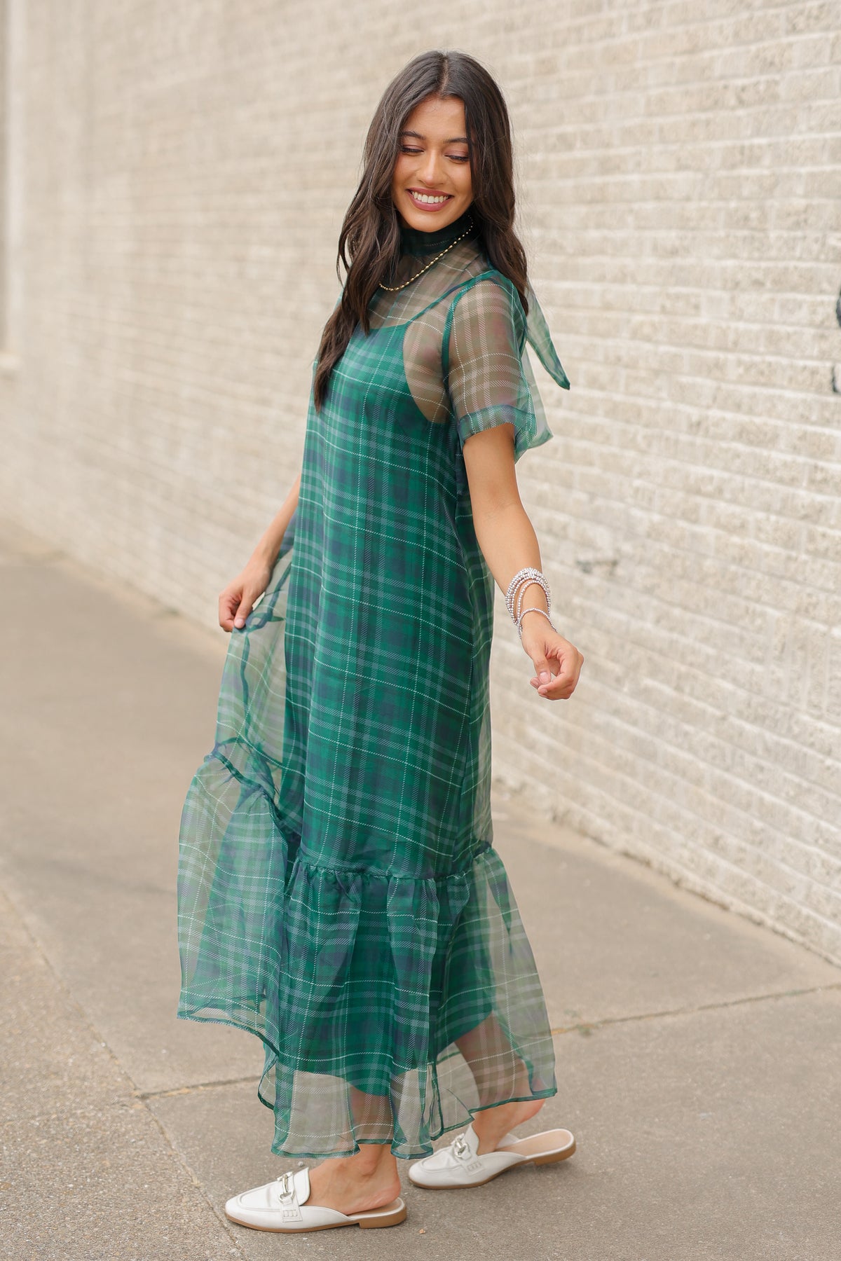 Model standing outside wearing the Glenhaven Plaid Ruffle Maxi Dress in emerald green and navy featuring a high neckline, sheer short sleeves, relaxed silhouette, and flowy ruffled hem. Southern House Clothing