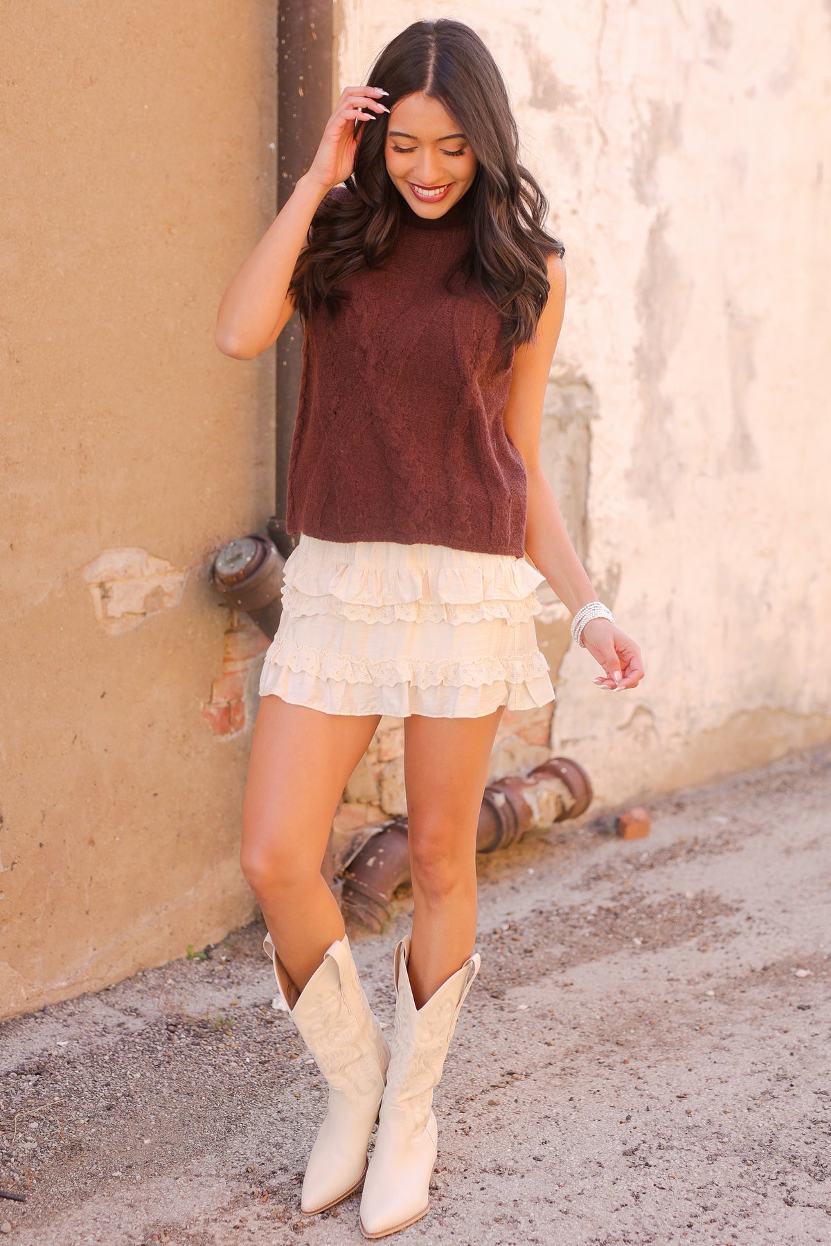 Woman wearing a maroon sleeveless top, white skirt, and beige cowboy boots against a textured wall.