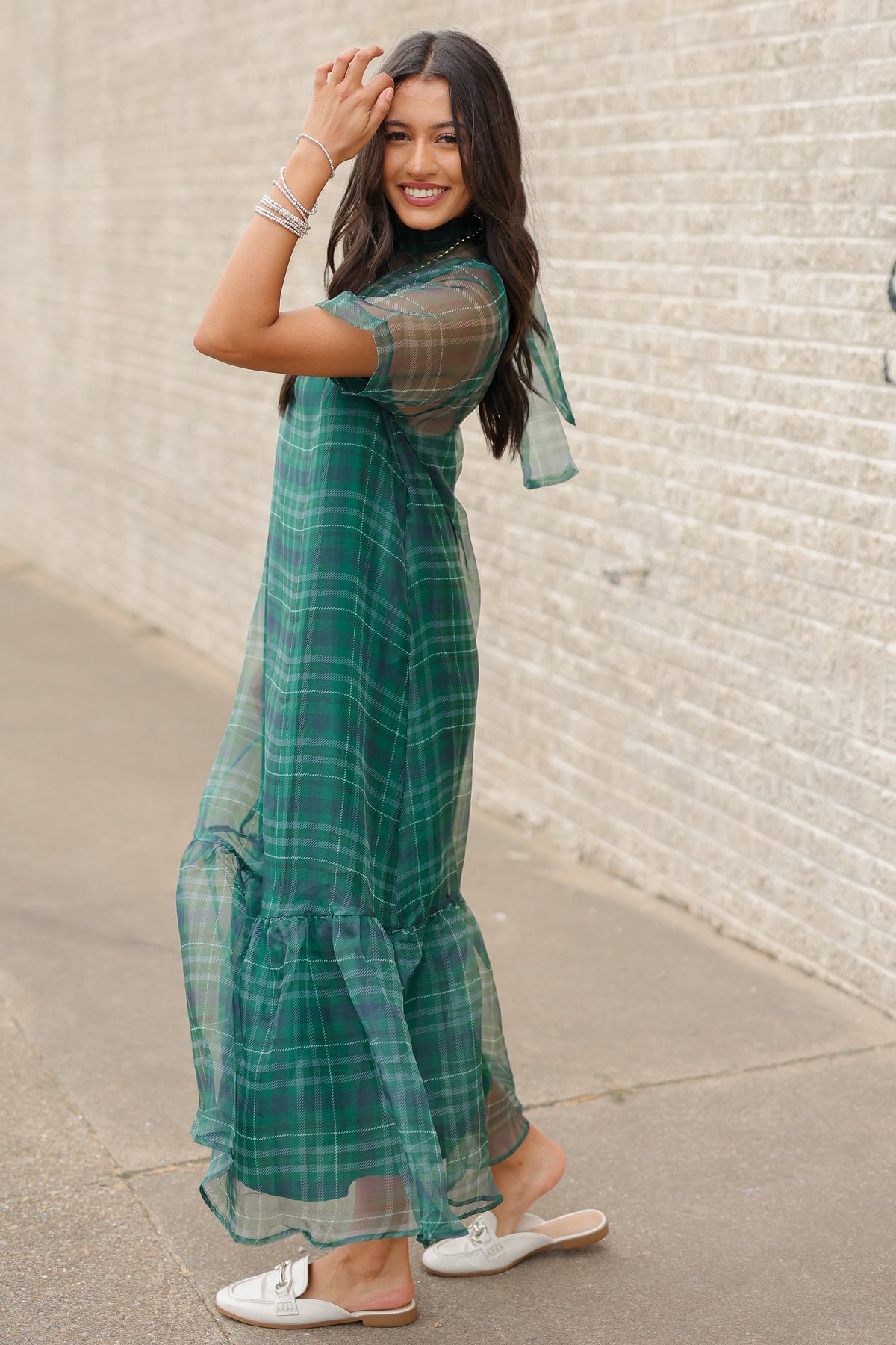 Model standing outside wearing the Glenhaven Plaid Ruffle Maxi Dress in emerald green and navy featuring a high neckline, sheer short sleeves, relaxed silhouette, and flowy ruffled hem. Southern House Clothing