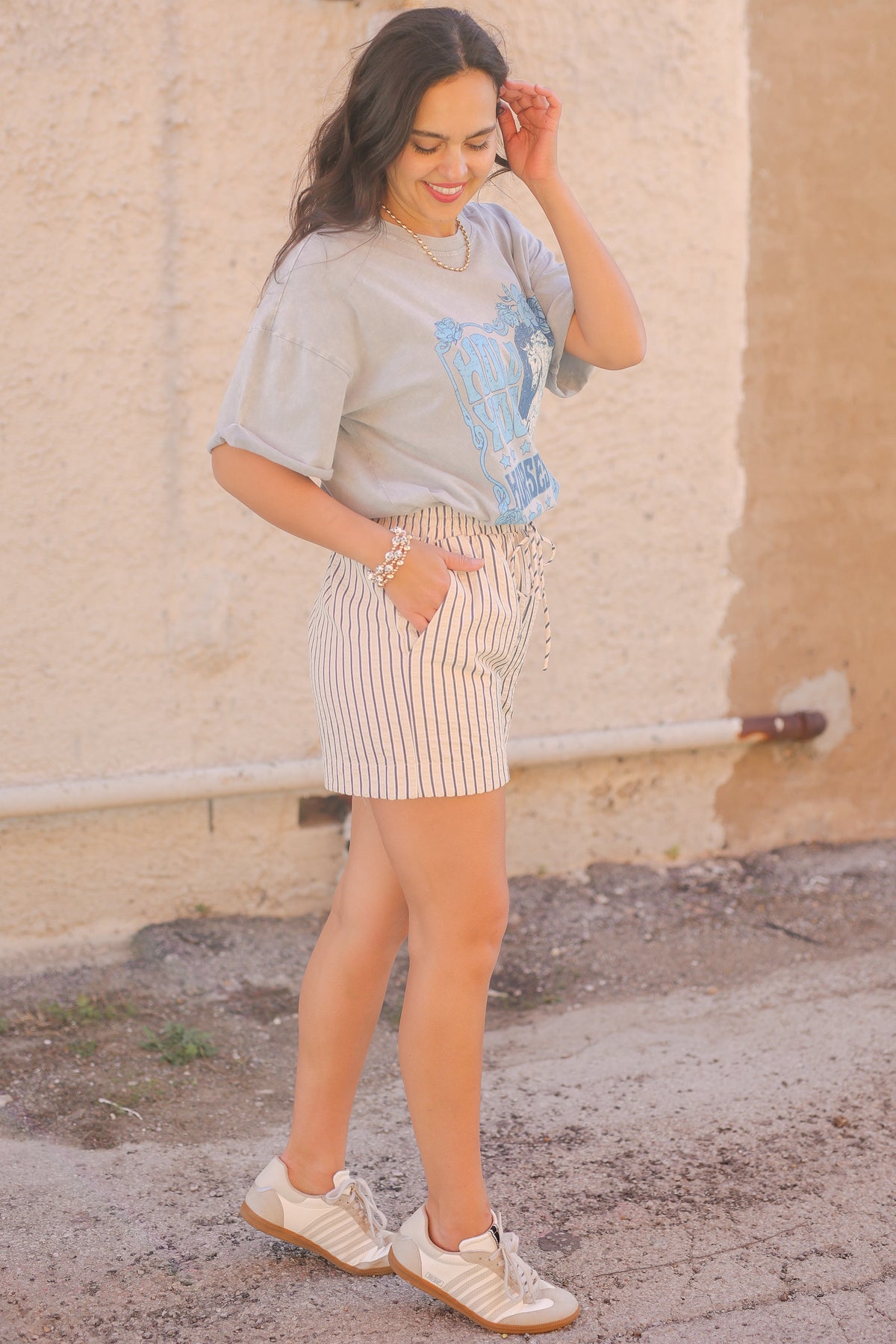 Woman wearing a light gray t-shirt with a colorful design and striped shorts, standing against a beige wall.