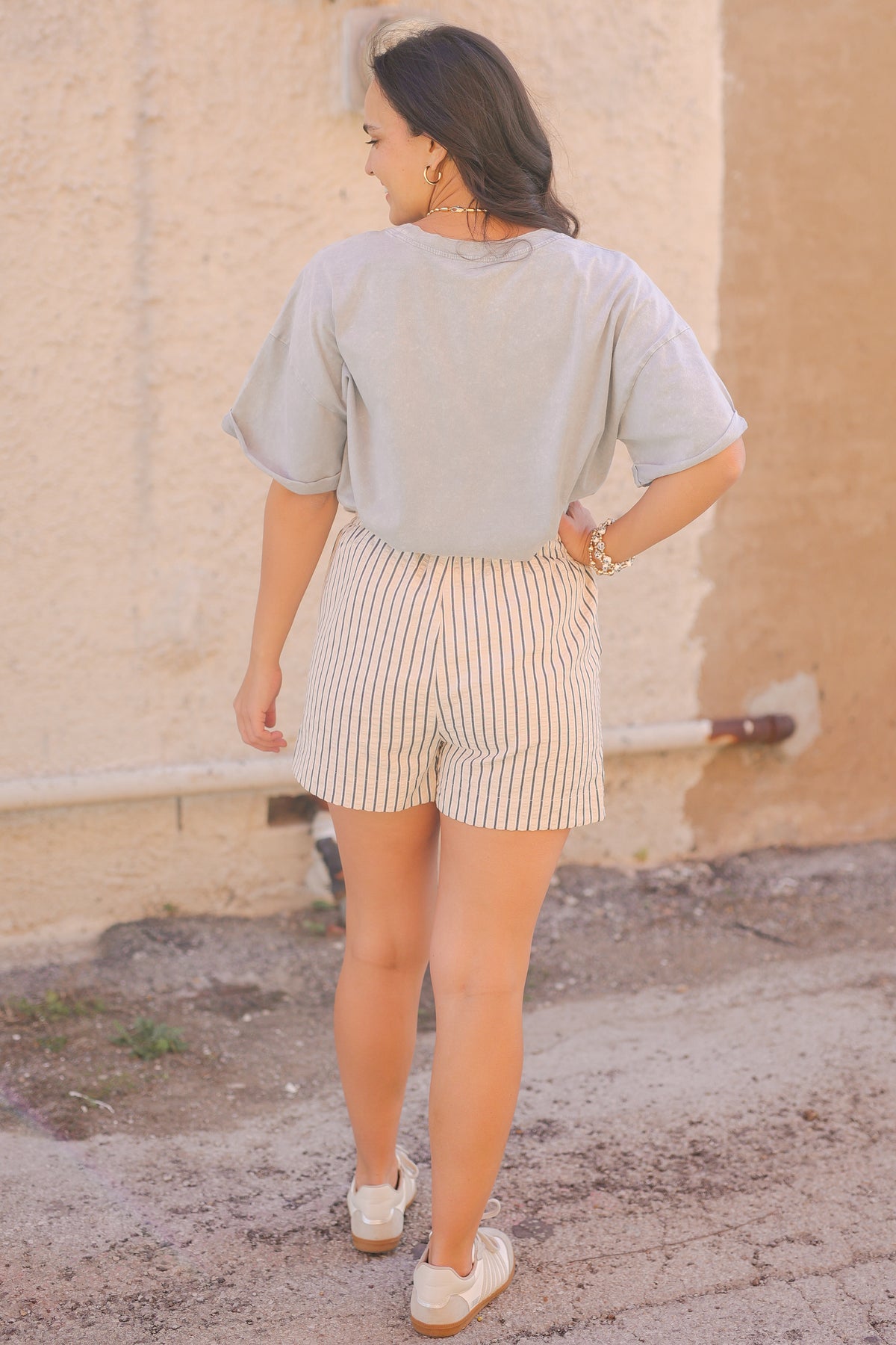 Woman wearing a light gray t-shirt and striped shorts standing against a beige wall.