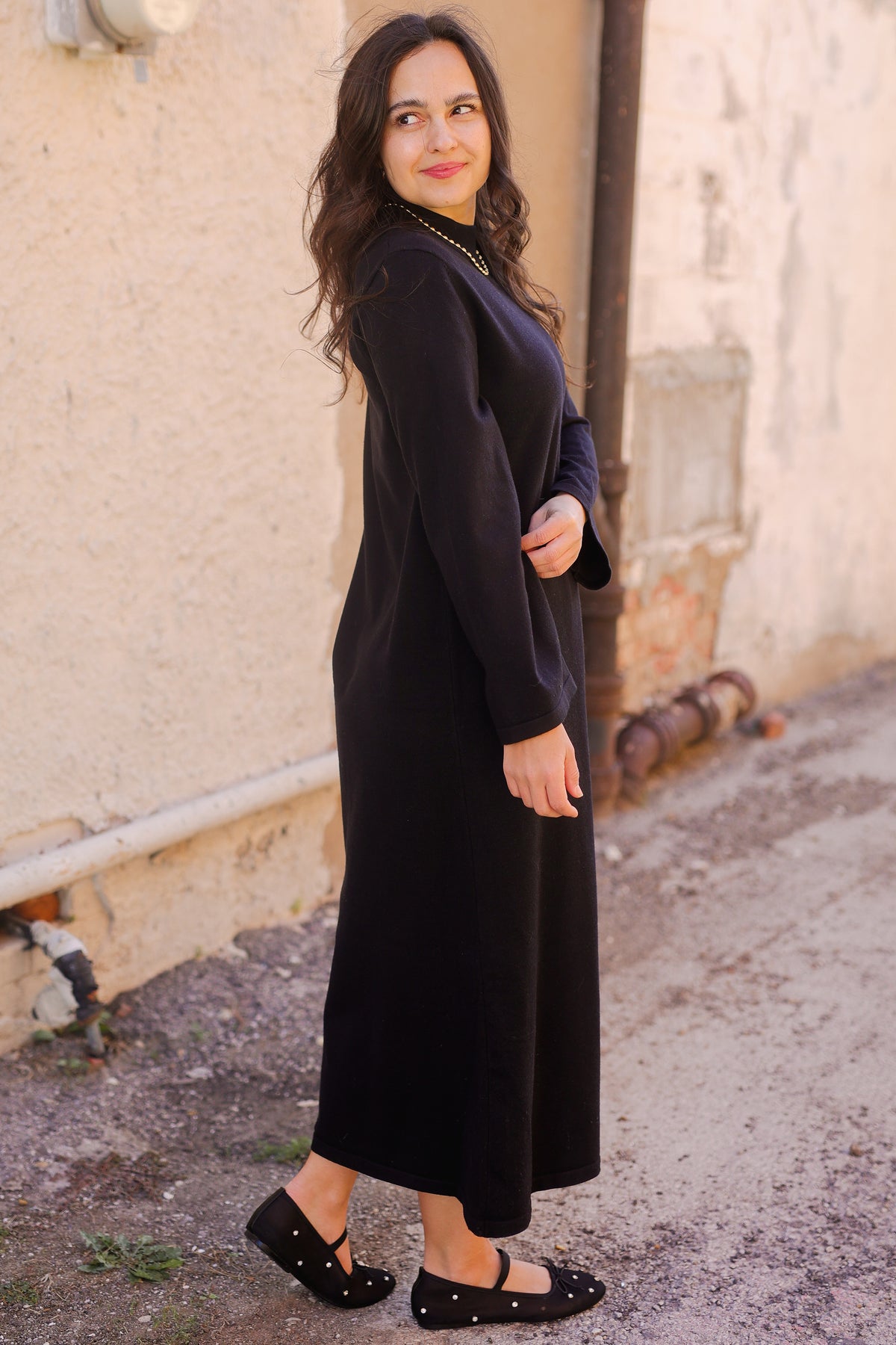 Woman wearing a long black dress standing against a textured wall.