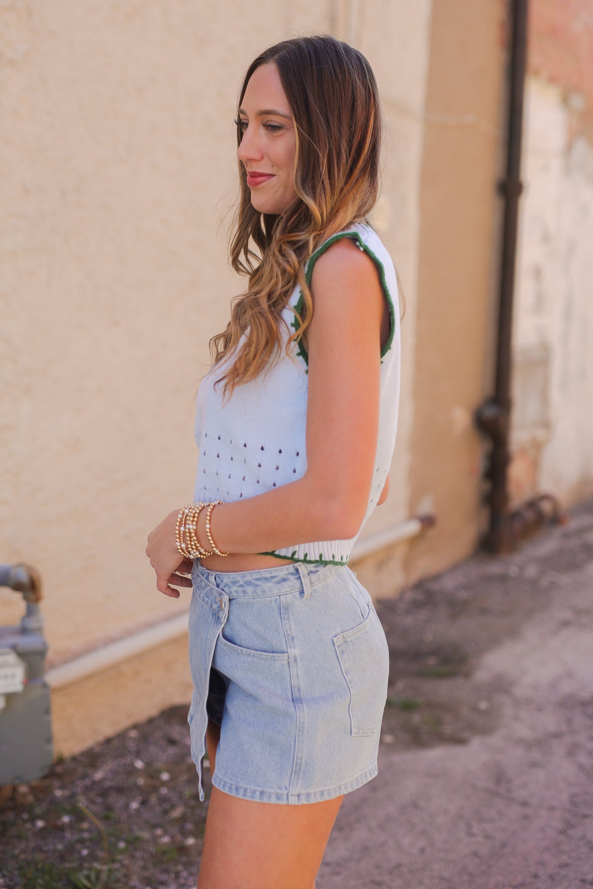Woman wearing a light blue sleeveless top and denim shorts standing against a beige wall.