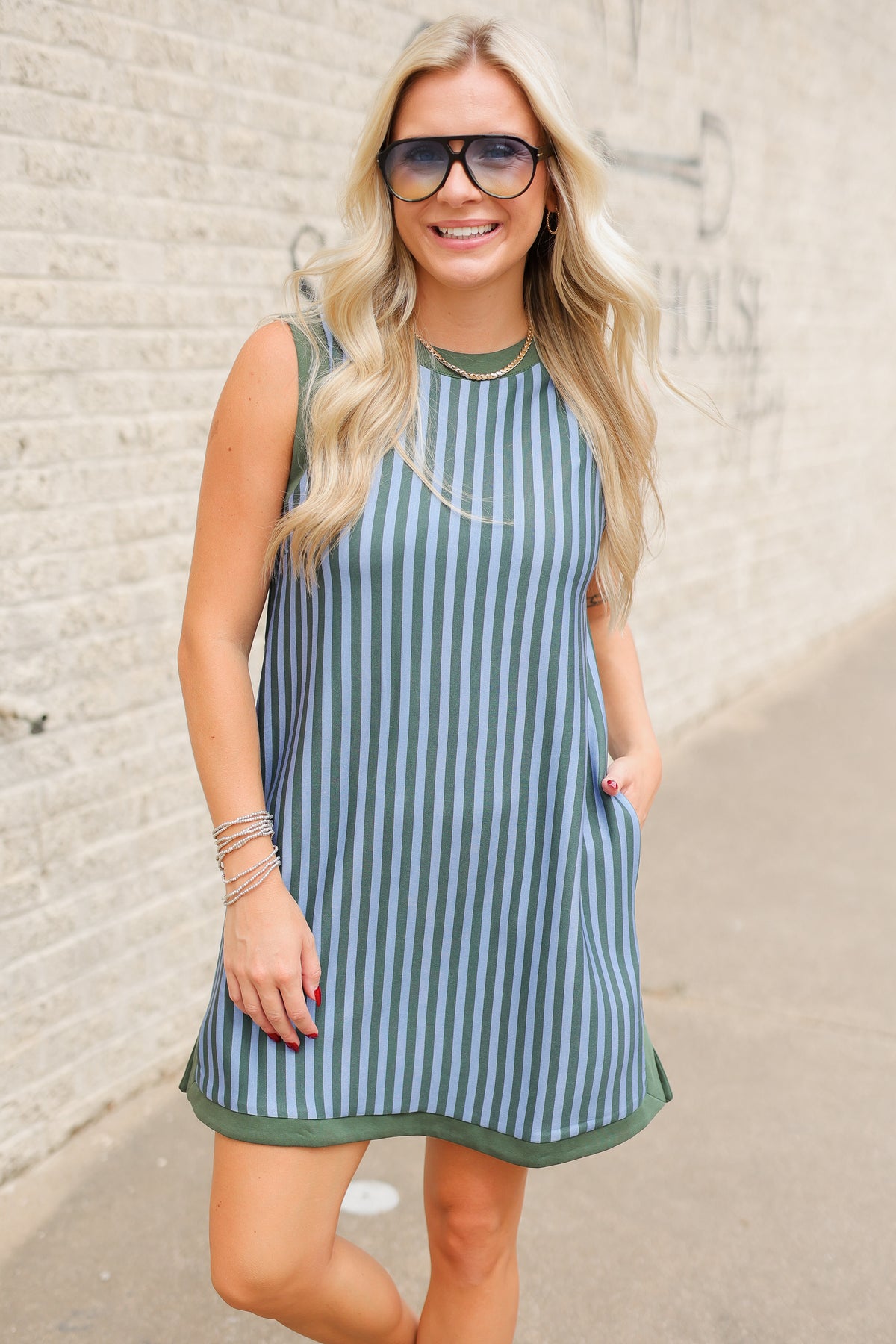 Blonde model wearing a green and blue striped dress and white flats standing outside. Southern House Clothing. Dress name is called the Aledo Lane Striped Dress