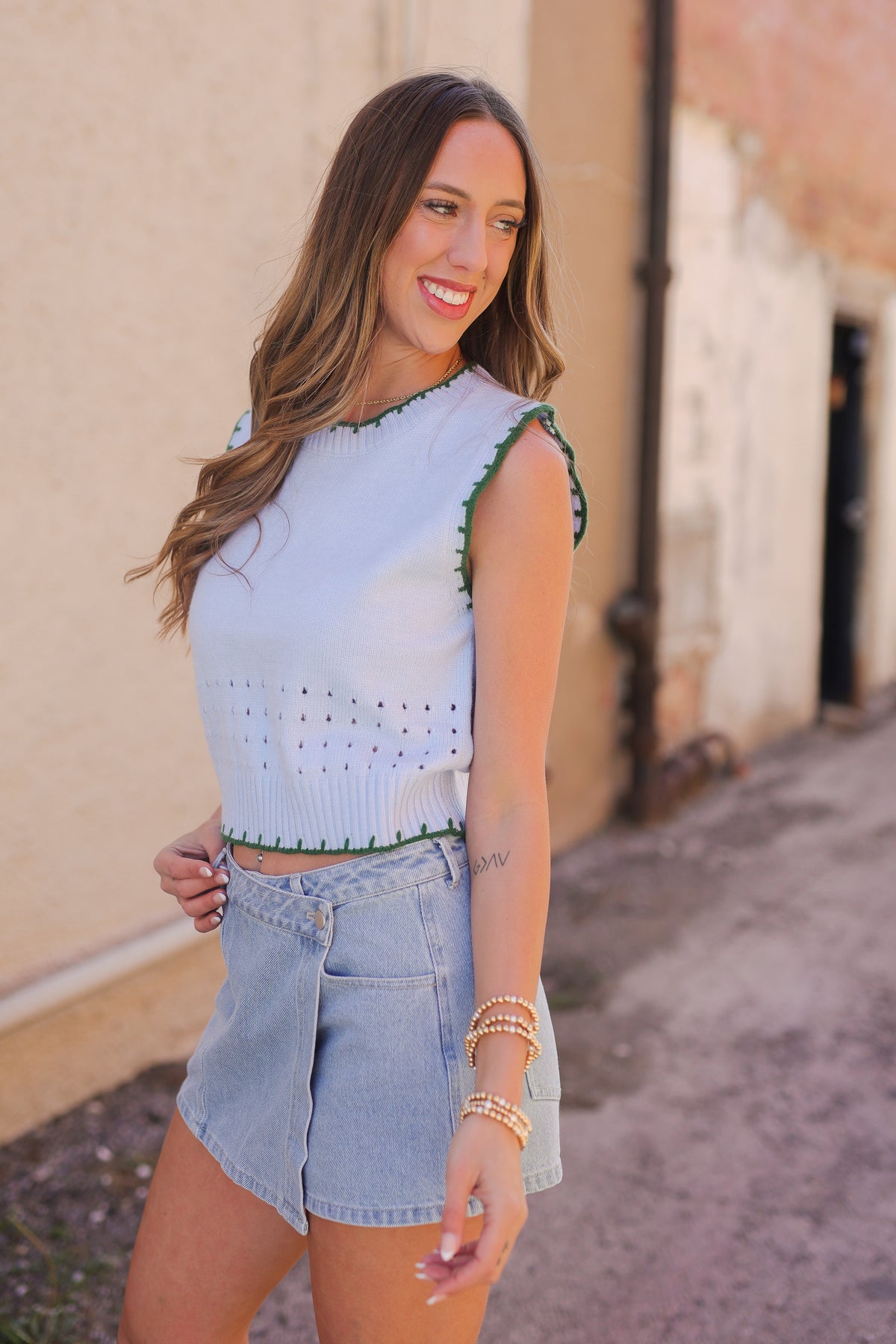 Woman wearing a light blue top with green accents and denim shorts standing against a beige wall.