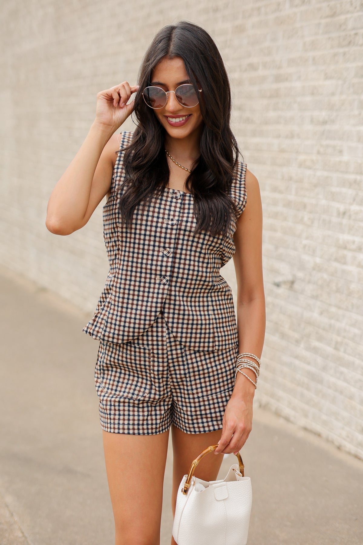 A woman stands outdoors against a light brick wall wearing a sleeveless brown and white gingham romper, brown knee-high boots, and holding a small white handbag.
