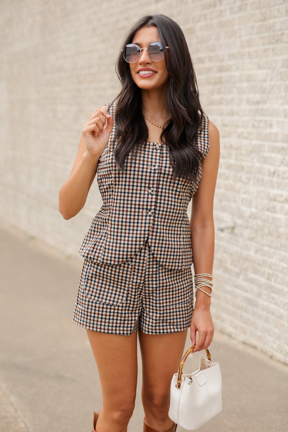 A woman wearing a brown, beige, and black gingham check two-piece set consisting of a sleeveless button-front top and matching shorts, styled with sunglasses, bracelets, and a white handbag against a light brick wall.