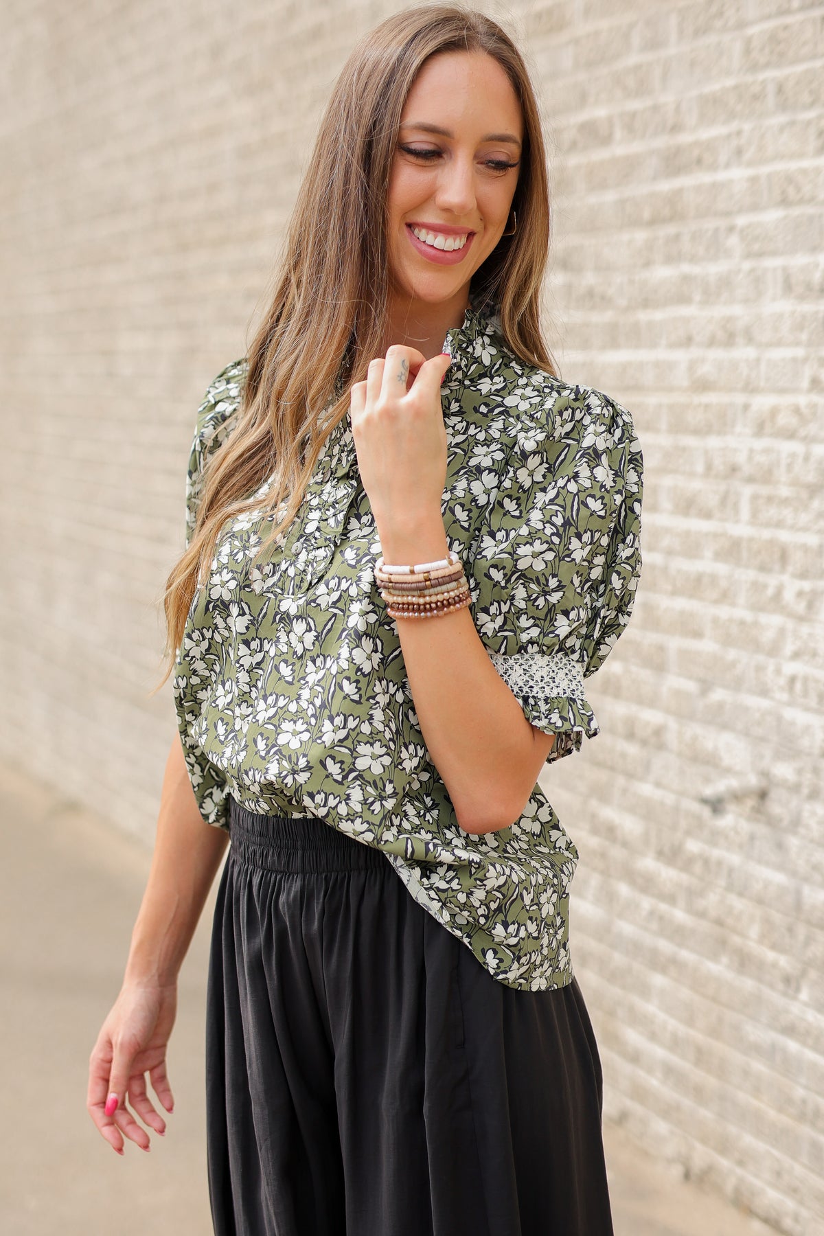 Blond model standing outside wearing a green blouse and black plants with bracelets and black ballet flats.