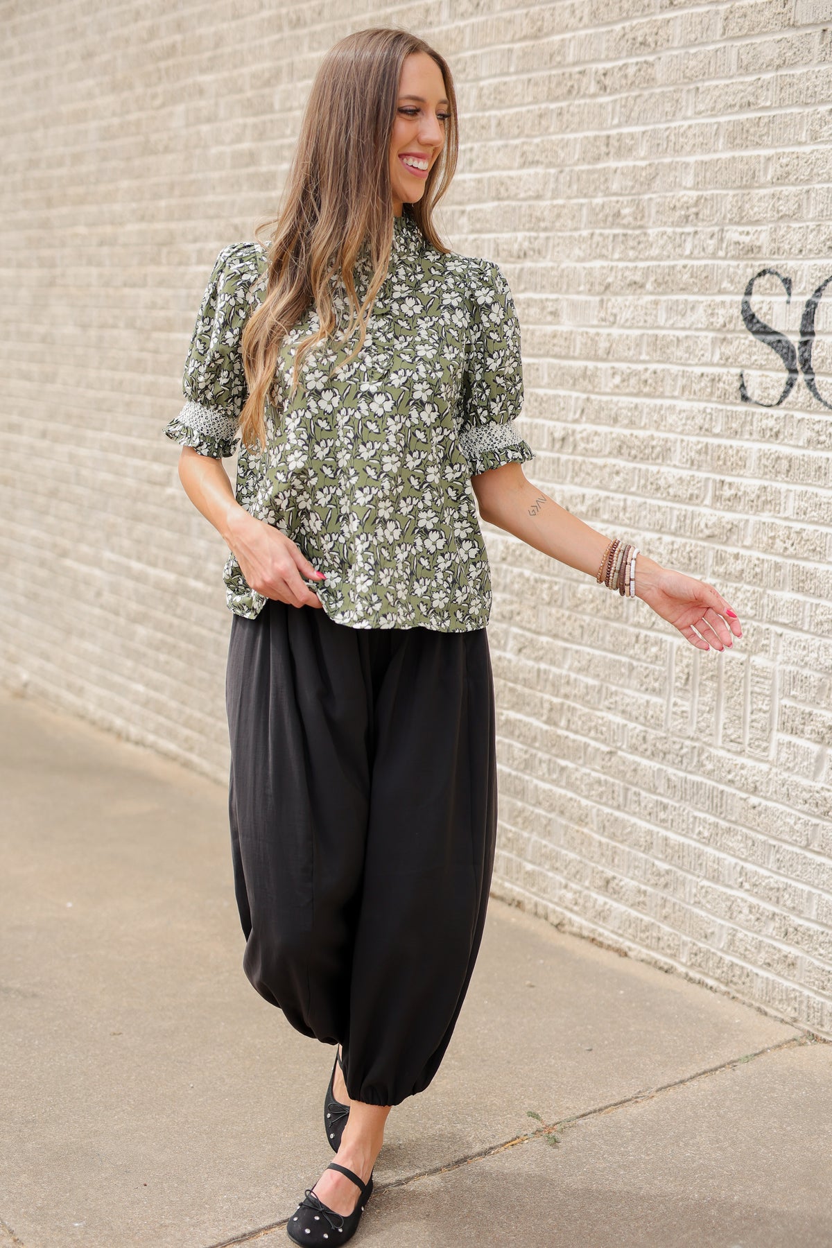 Blond model standing outside wearing a green blouse and black plants with bracelets and black ballet flats.