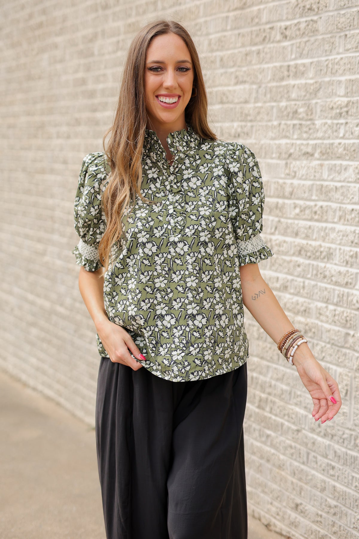 Blond model standing outside wearing a green blouse and black plants with bracelets and black ballet flats.