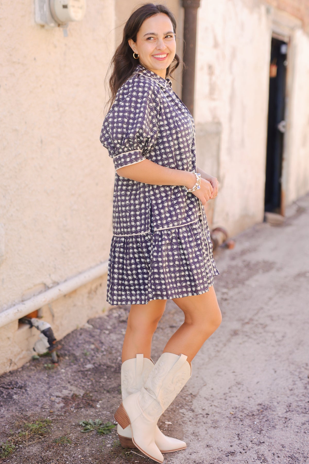 Woman wearing a blue checkered dress and white cowboy boots standing on a street.