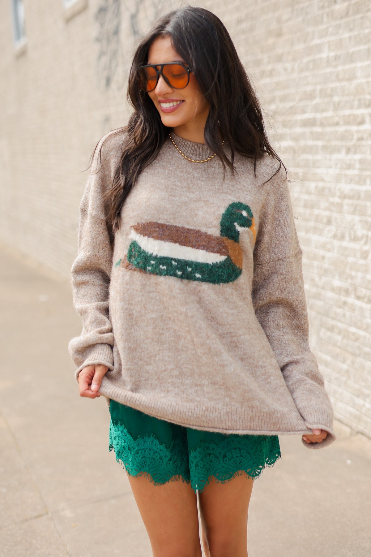Brunette model wearing a mallard print knit sweater and green lace shorts with white cowboy boots standing outside. Southern House Clothing