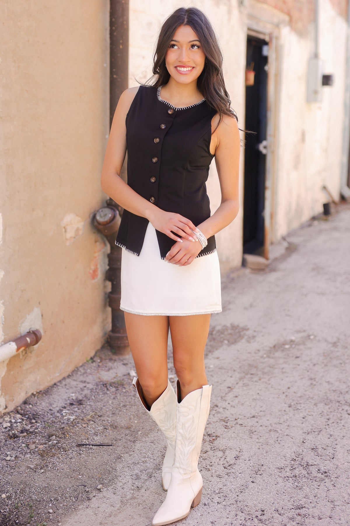 Woman wearing a black vest, white dress, and white cowboy boots standing on a street.
