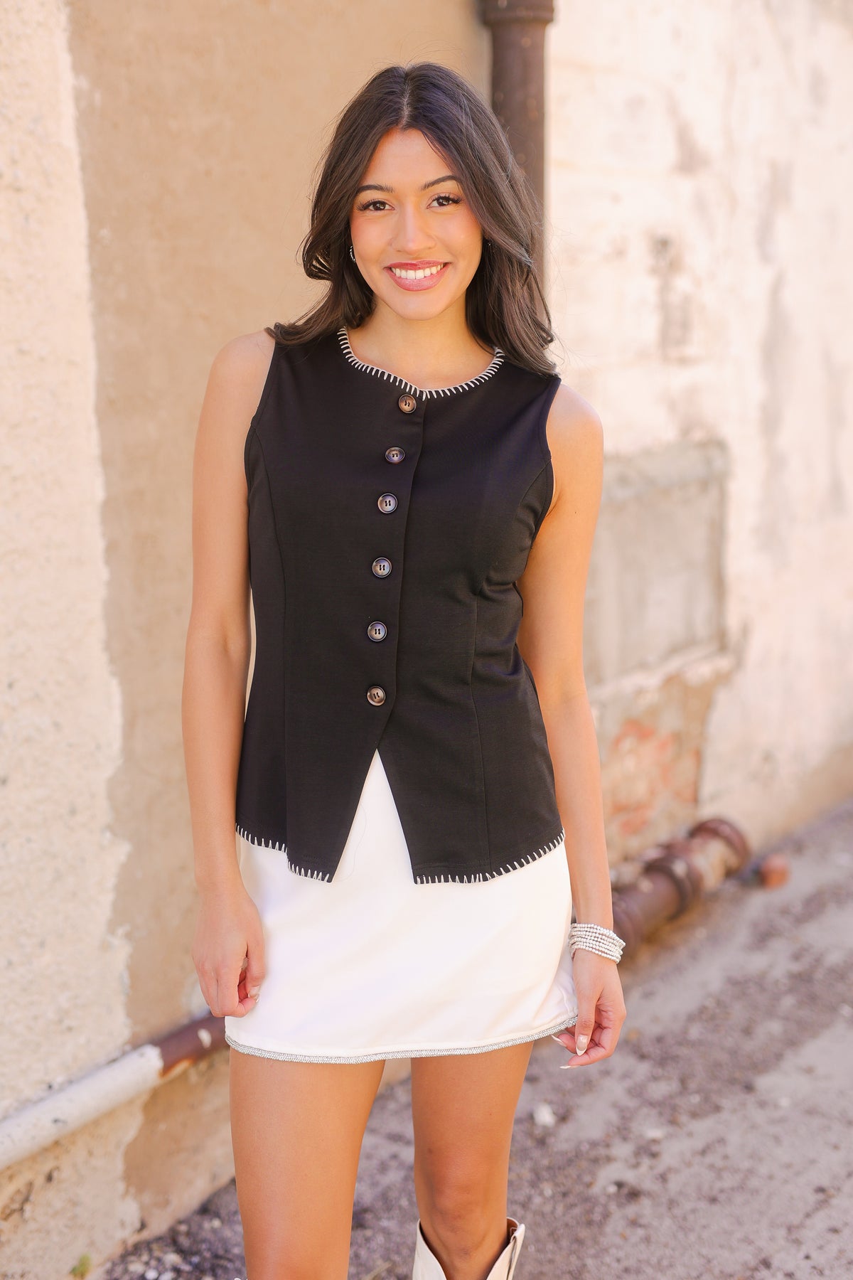 Woman wearing a black vest over a white dress standing against a beige wall.