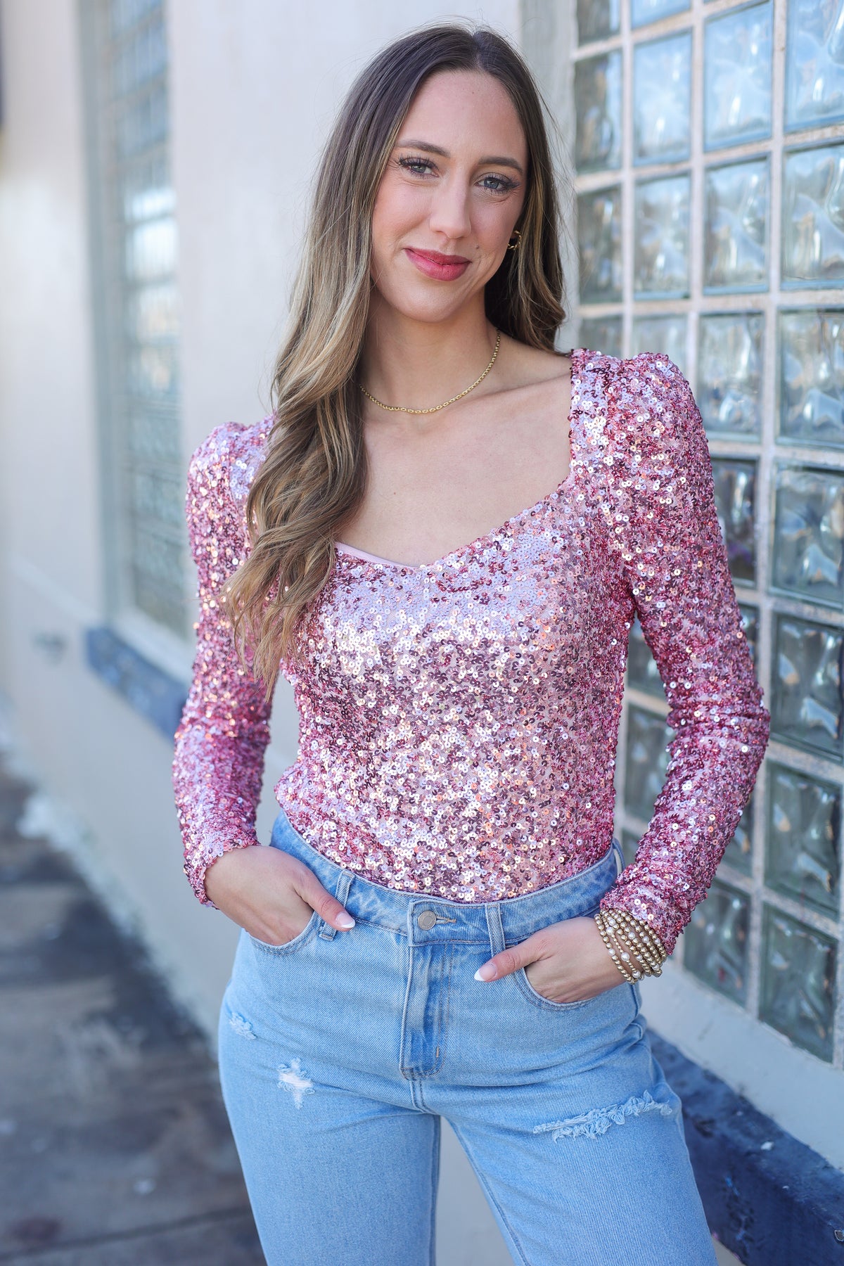 Woman wearing a pink sequin top and blue jeans standing in front of a glass block wall.