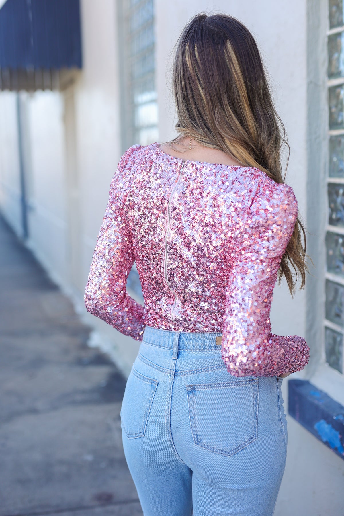 Person wearing a pink sequin top and light blue jeans standing against a white wall.