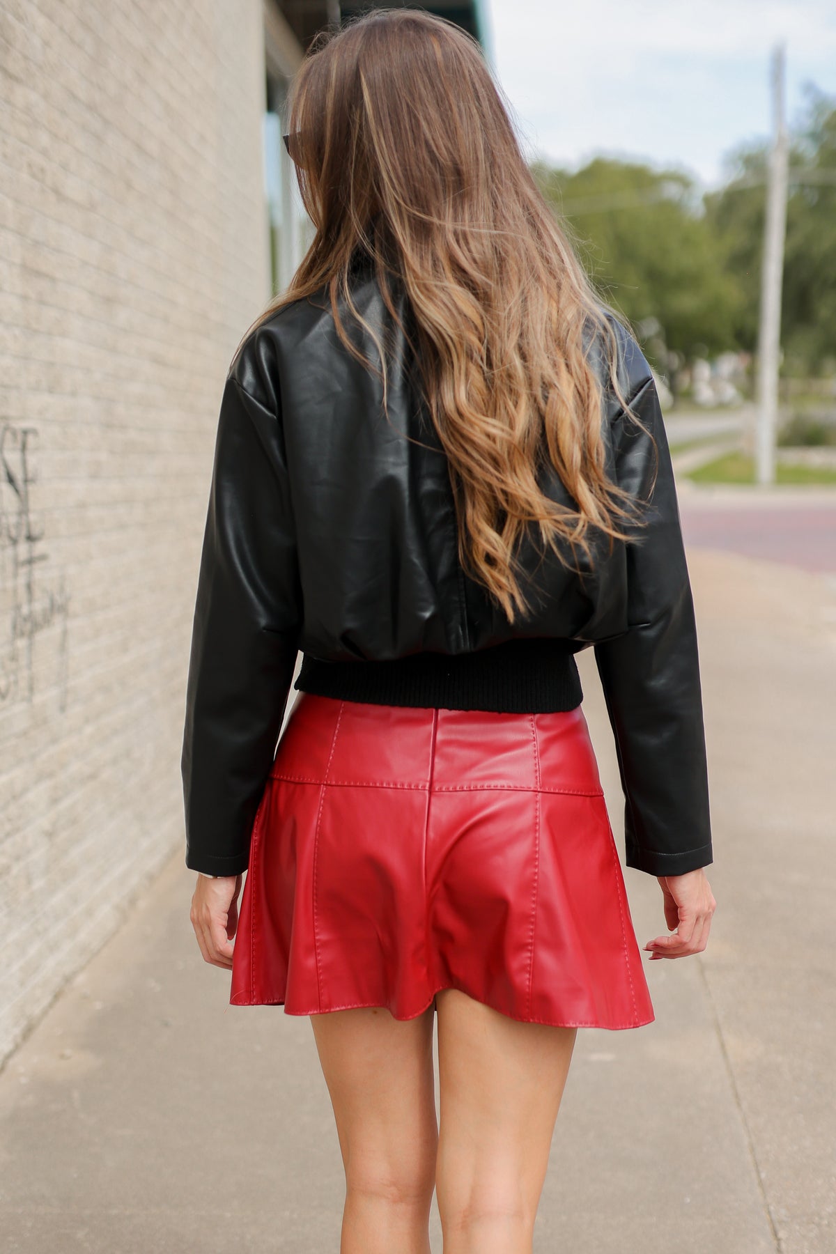 Behind view of a model wearing a red skirt and leather crop jacket. Standing outside. Southern House Clothing
