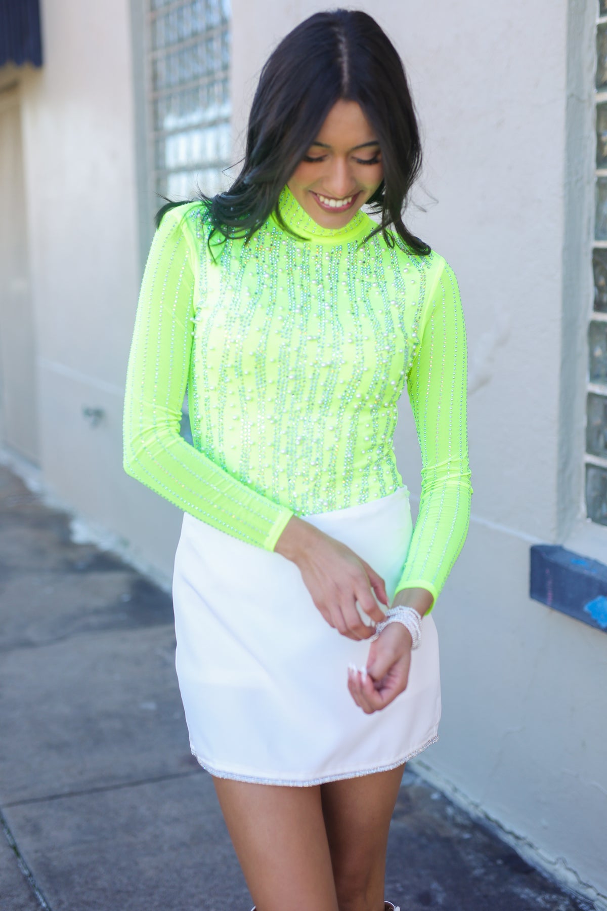 Woman wearing a bright green top and white skirt standing against a light-colored wall.