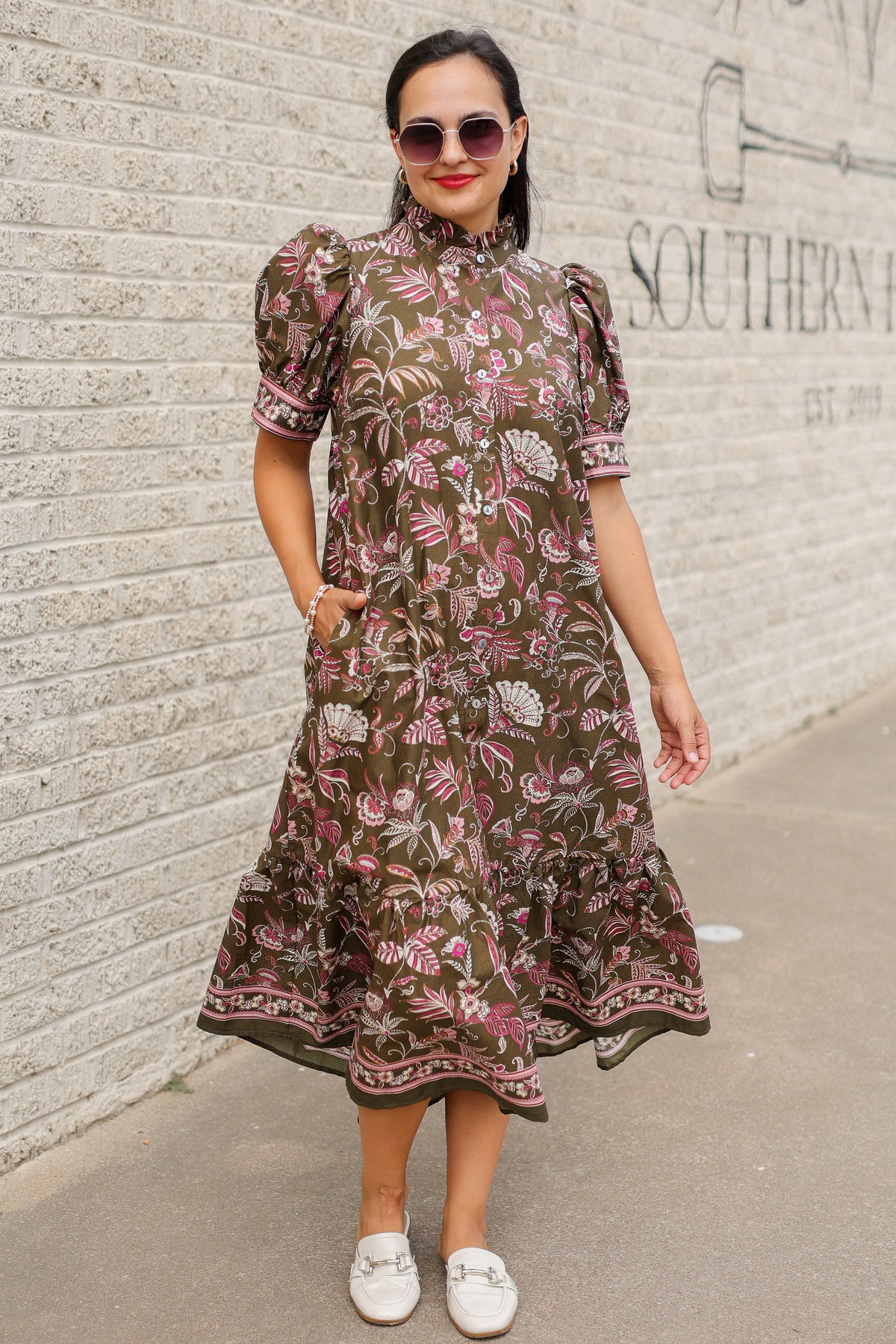 Brunette model wearing an olive dress with patterns standing outside. Southern House Clothing