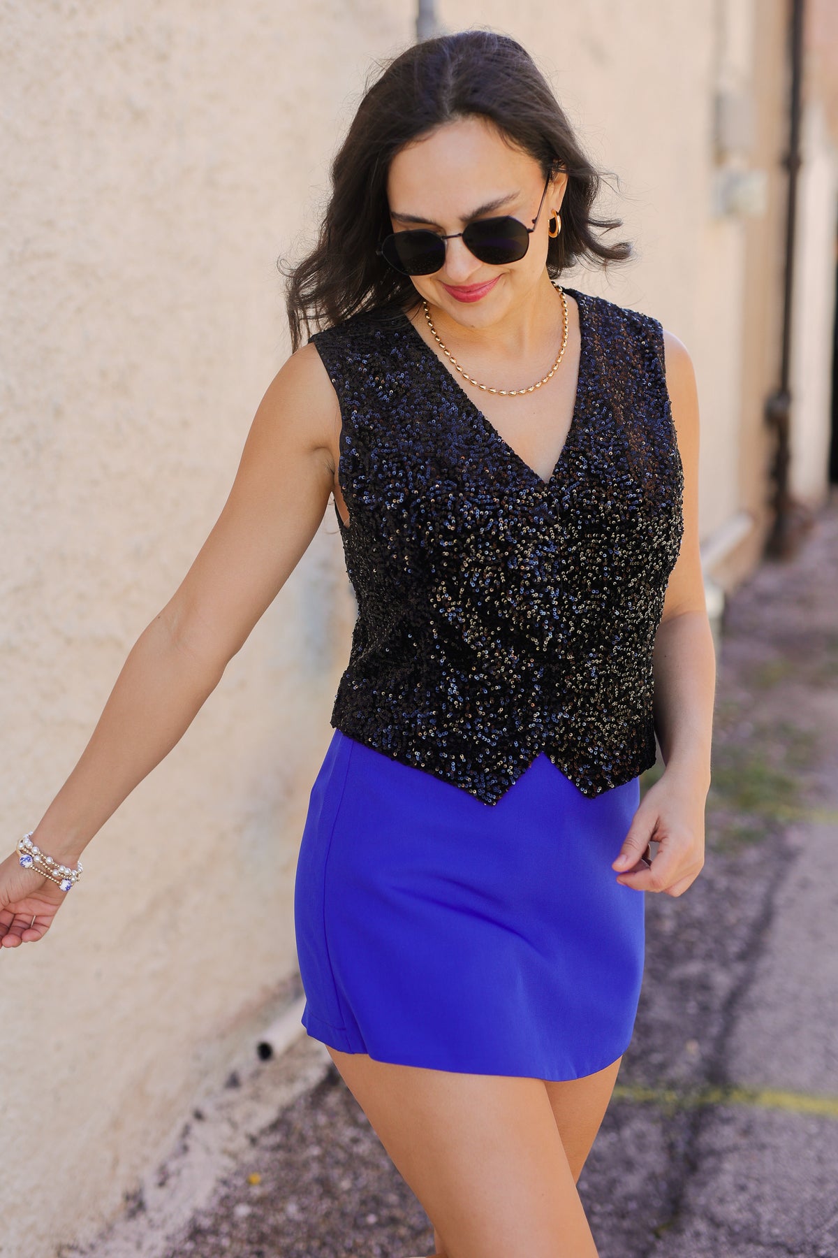 Woman wearing a black sequin top and blue skirt against a beige wall.