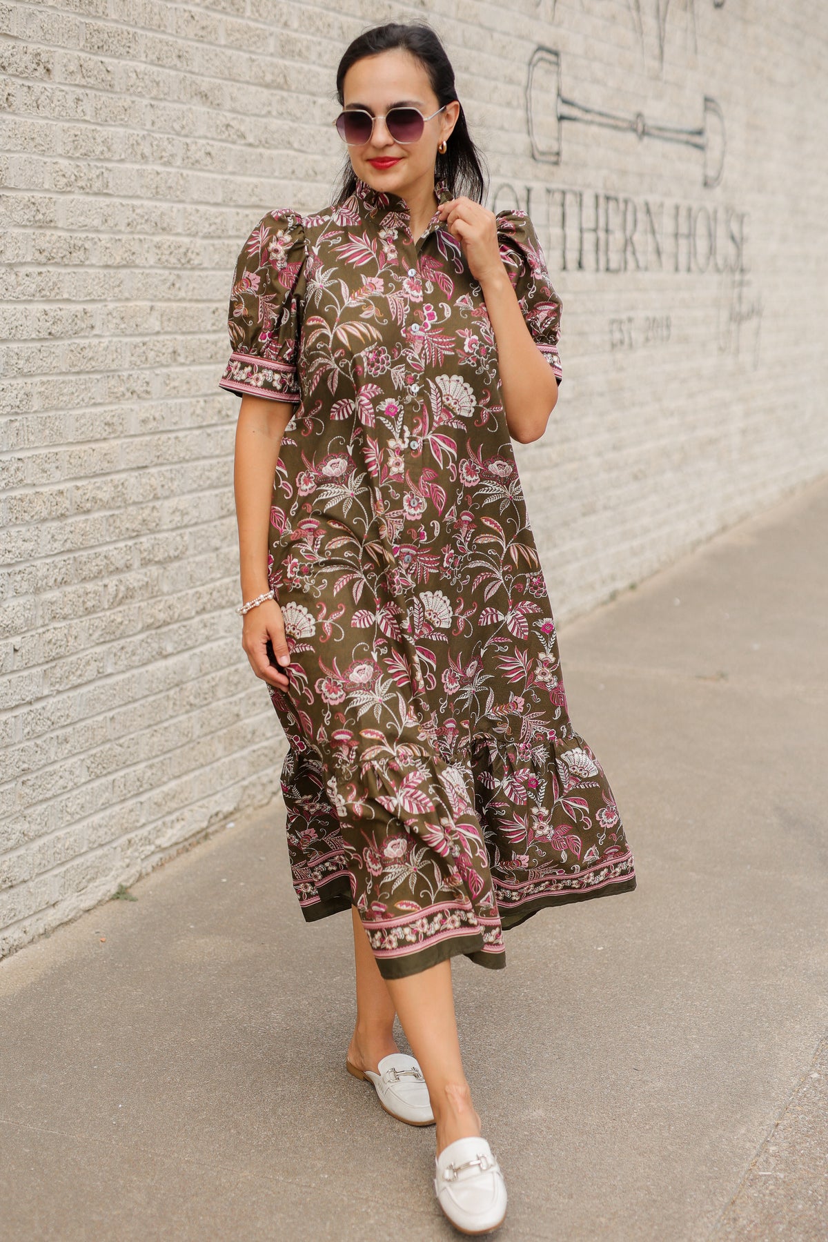Brunette model wearing an olive dress with patterns standing outside. Southern House Clothing