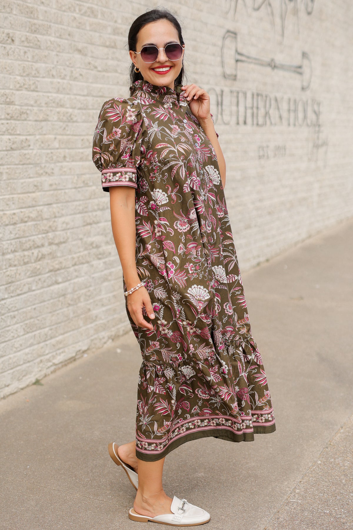Brunette model wearing an olive dress with patterns standing outside. Southern House Clothing