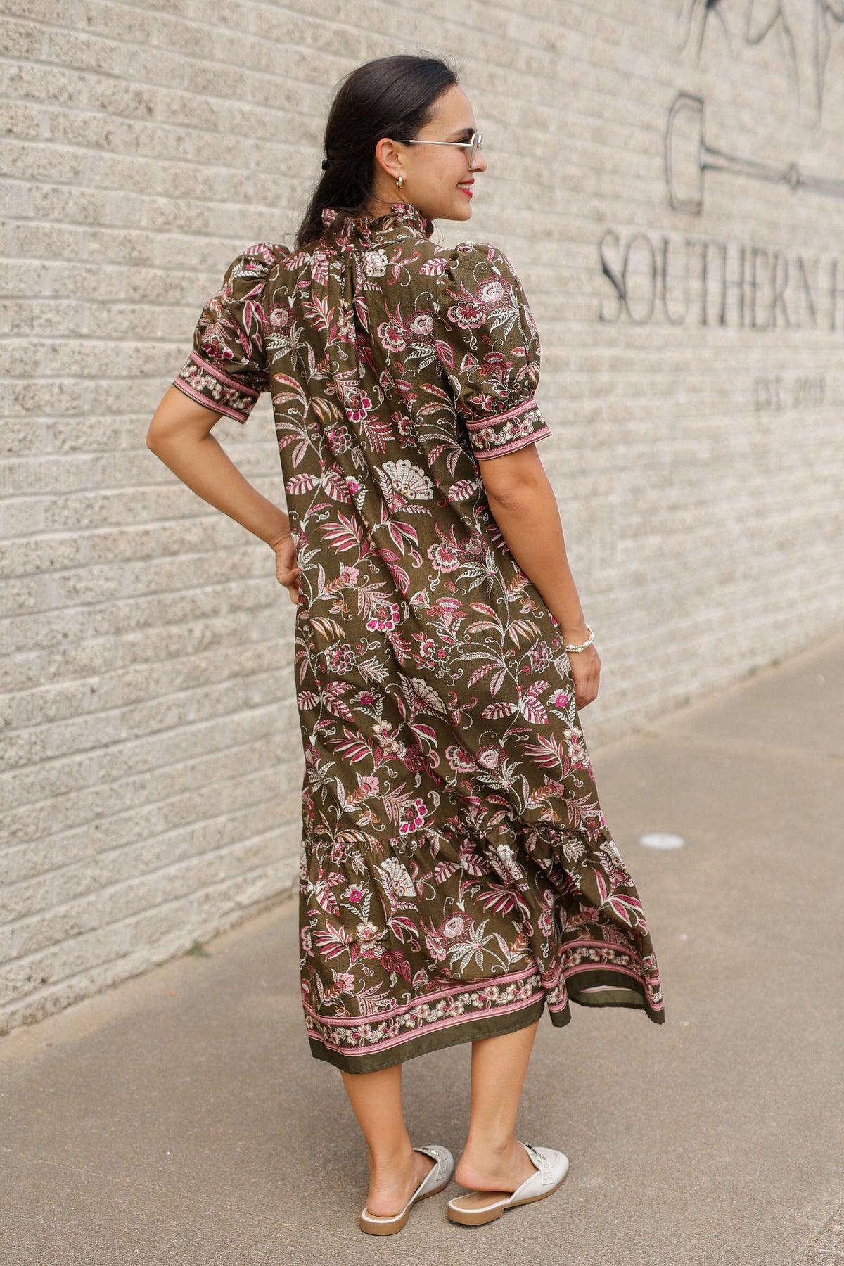Brunette model wearing an olive dress with patterns standing outside. Southern House Clothing