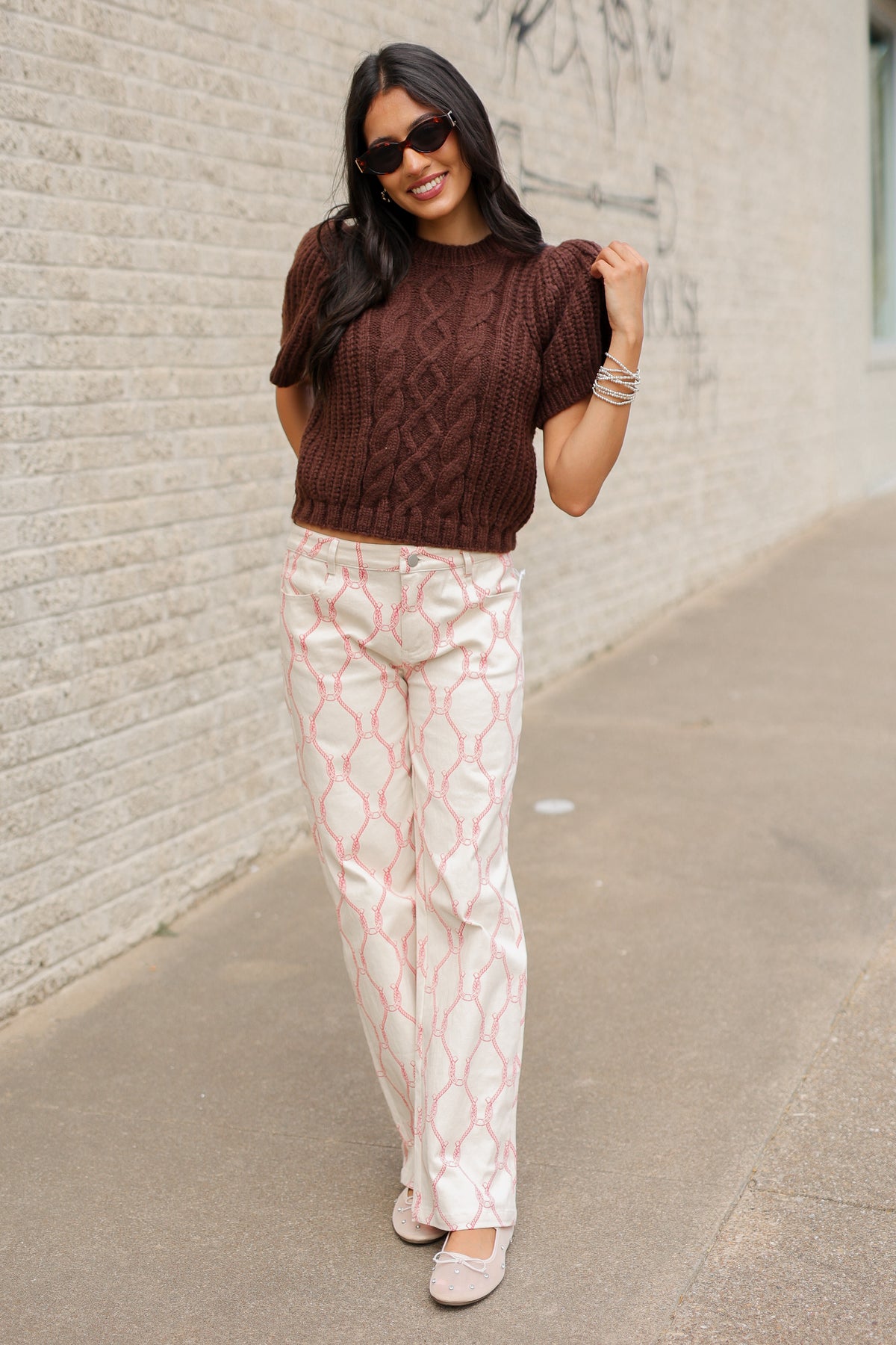 Brunette model wearing the Huxley Cable Knit Sweater Top in Dark Chocolate featuring a textured cable knit pattern, ribbed crew neckline, and short puff sleeves, paired with the Equestrian Charm Horseshoe Print Pants in Ivory and Pink styled, featuring an allover pink rope and horseshoe design. Southern House Clothing