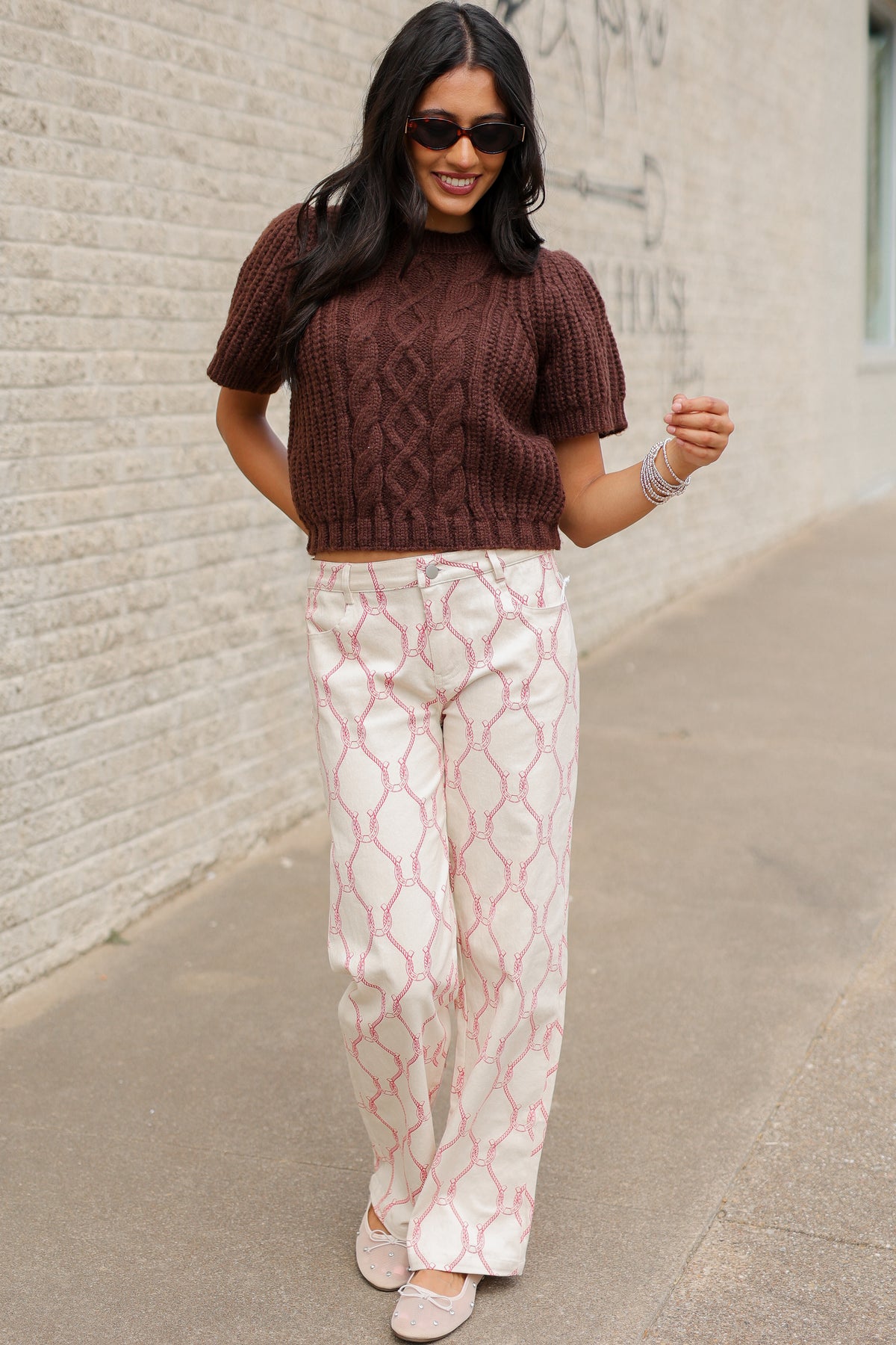 Brunette model wearing the Huxley Cable Knit Sweater Top in Dark Chocolate featuring a textured cable knit pattern, ribbed crew neckline, and short puff sleeves, paired with the Equestrian Charm Horseshoe Print Pants in Ivory and Pink styled, featuring an allover pink rope and horseshoe design. Southern House Clothing