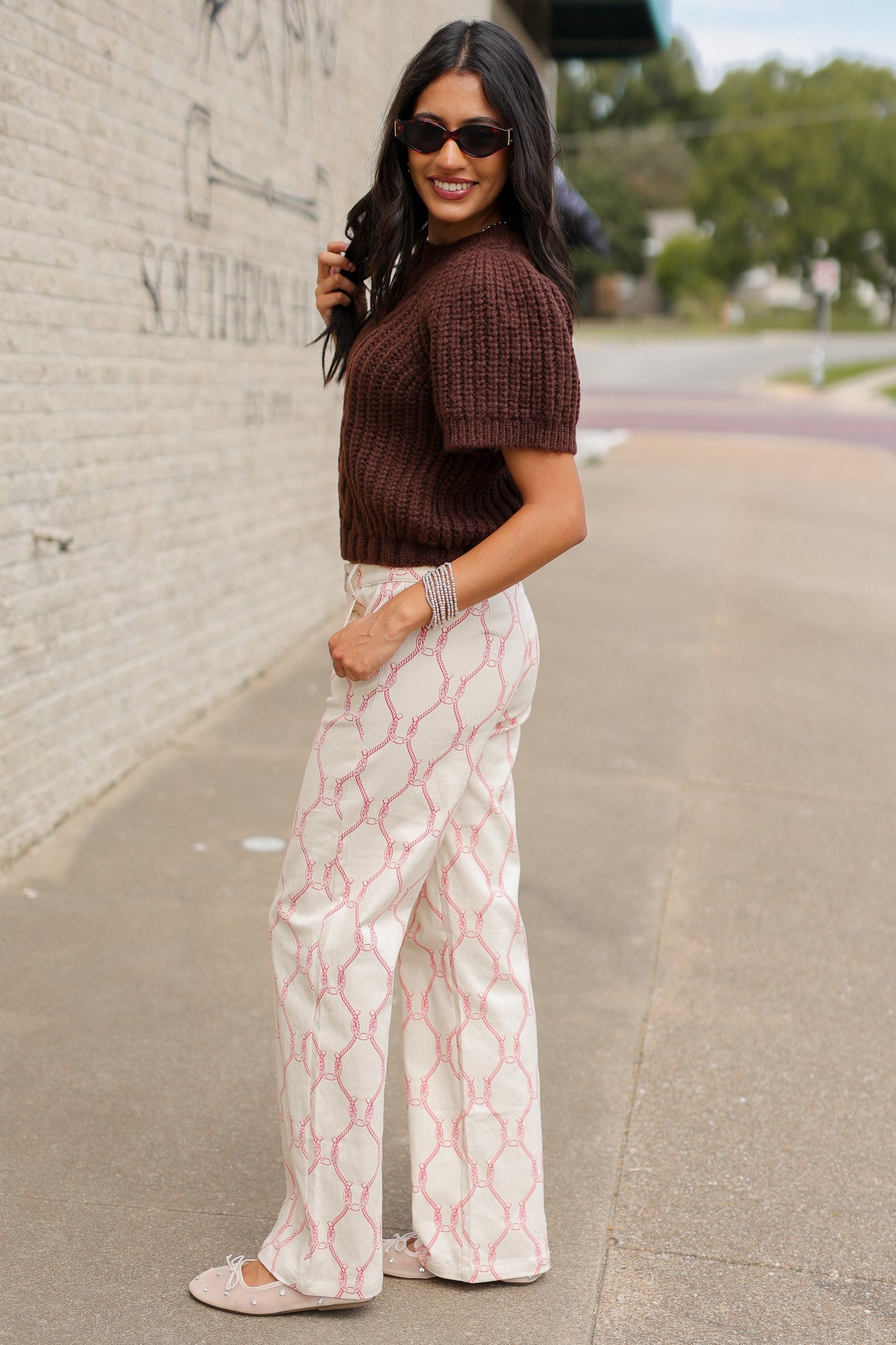 Brunette model wearing the Huxley Cable Knit Sweater Top in Dark Chocolate featuring a textured cable knit pattern, ribbed crew neckline, and short puff sleeves, paired with the Equestrian Charm Horseshoe Print Pants in Ivory and Pink styled, featuring an allover pink rope and horseshoe design. Southern House Clothing