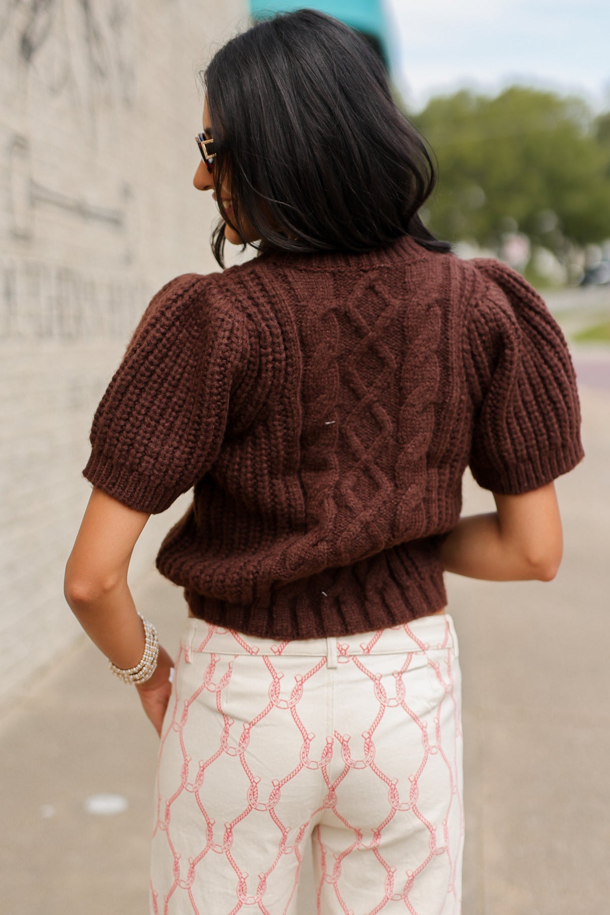 Behind view of a Brunette model wearing the Huxley Cable Knit Sweater Top in Dark Chocolate featuring a textured cable knit pattern, ribbed crew neckline, and short puff sleeves, paired with the Equestrian Charm Horseshoe Print Pants in Ivory and Pink styled, featuring an allover pink rope and horseshoe design. Southern House Clothing