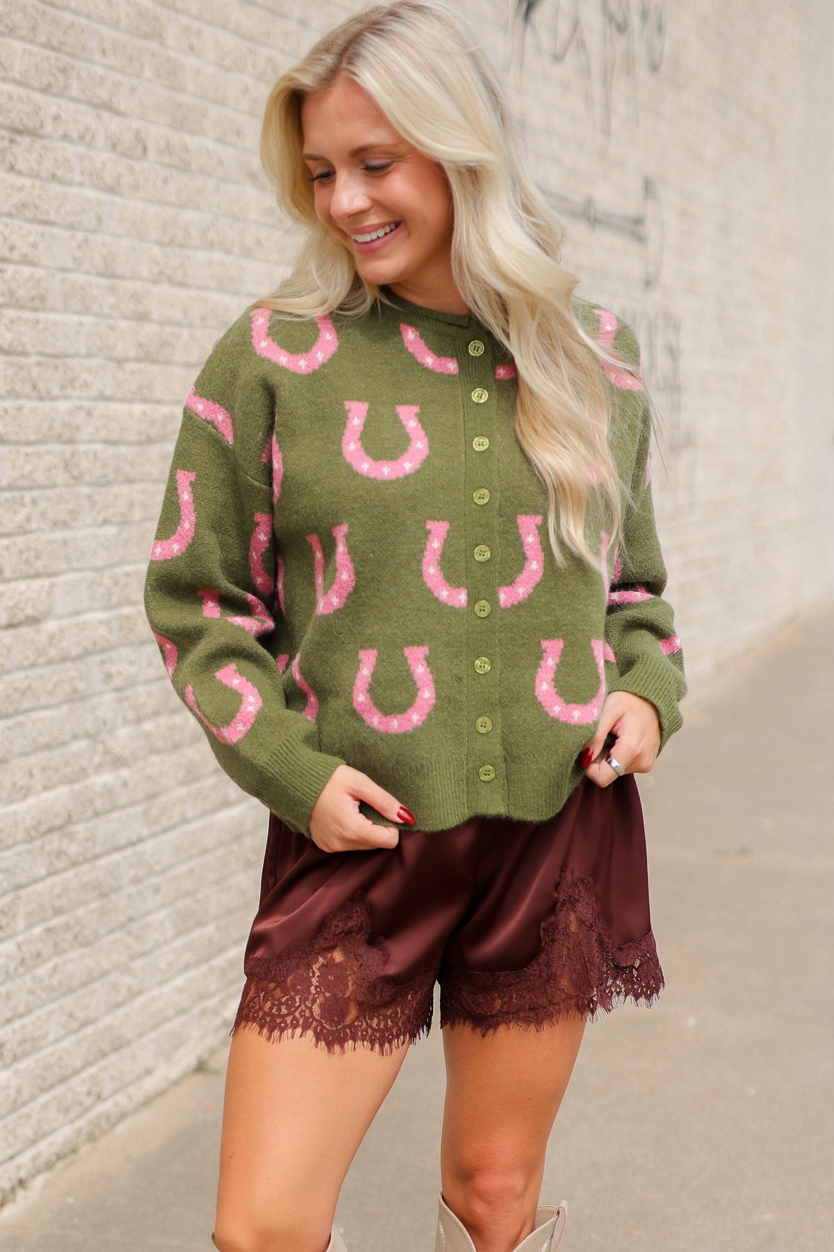 Model wearing the Roscoe Lucky Horseshoe Cardigan in olive green with oversized pink horseshoe designs, olive button-front closure, and ribbed cuffs, styled with Blair Lace Shorts chocolate brown lace-trim shorts. Southern House Clothing