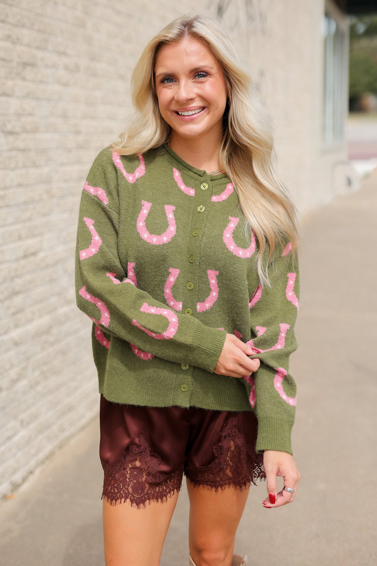 Model wearing the Roscoe Lucky Horseshoe Cardigan in olive green with oversized pink horseshoe designs, olive button-front closure, and ribbed cuffs, styled with Blair Lace Shorts chocolate brown lace-trim shorts. Southern House Clothing