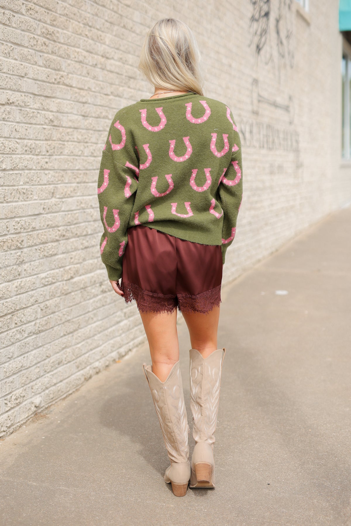 Model wearing the Roscoe Lucky Horseshoe Cardigan in olive green with oversized pink horseshoe designs, olive button-front closure, and ribbed cuffs, styled with Blair Lace Shorts chocolate brown lace-trim shorts. Southern House Clothing