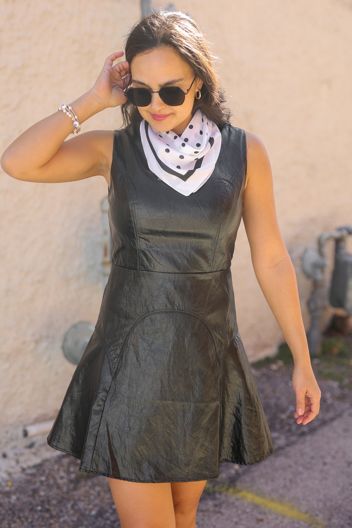 Woman wearing a black leather dress with sunglasses and a scarf against a beige wall.