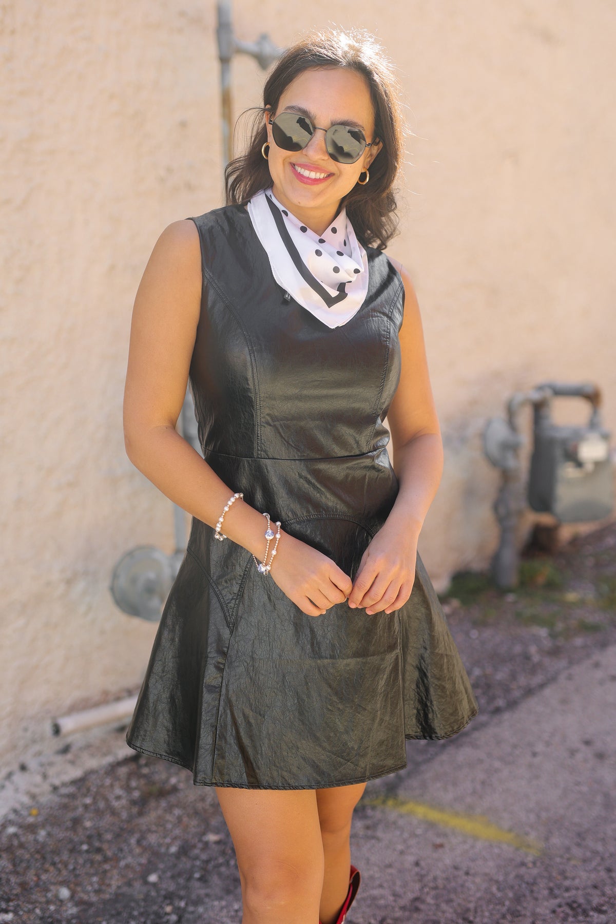 Woman wearing a black leather dress with sunglasses against a beige wall.