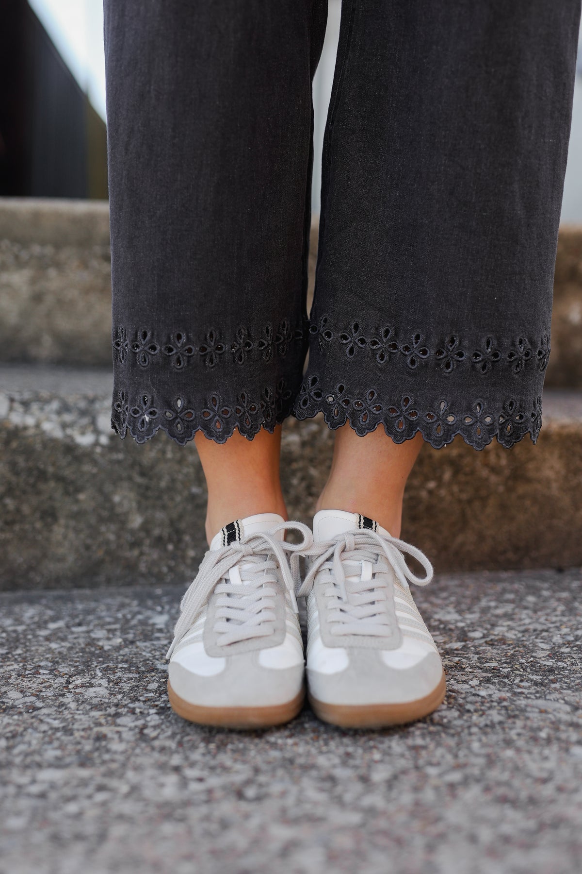 Person wearing black pants with lace trim and white sneakers on a concrete surface.