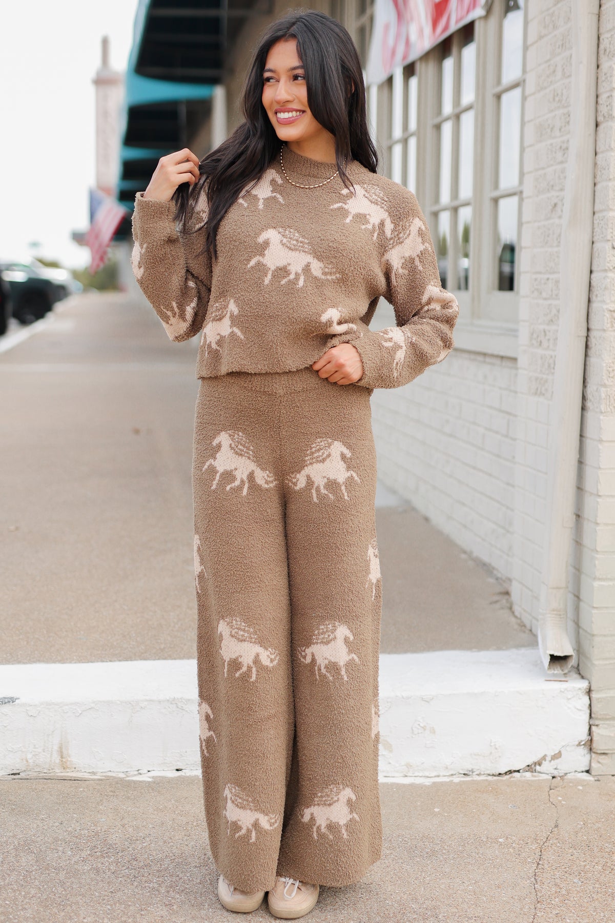 Brunette model wearing Bridlebrook Running Horse Sweater in Mocha featuring a fuzzy textured knit, cream running horse pattern, crew neckline, and long sleeves. it is paired with the Bridlebrook Running Horse Pants with the same pattern to make a set. Southern House Clothing