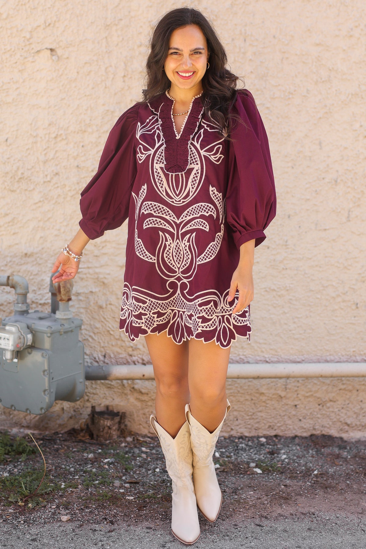Woman wearing a burgundy embroidered dress with white boots standing against a beige wall.