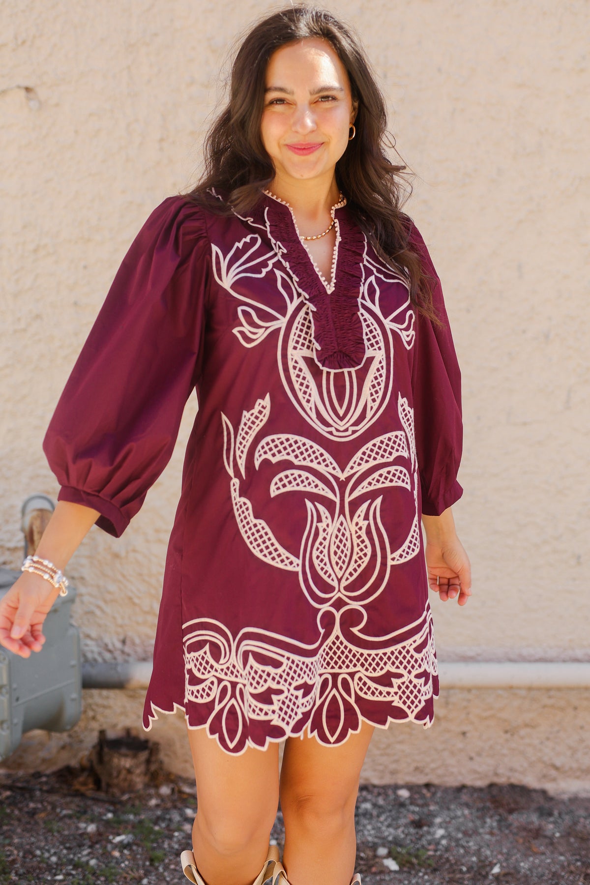 Woman wearing a burgundy dress with white embroidery against a beige wall.
